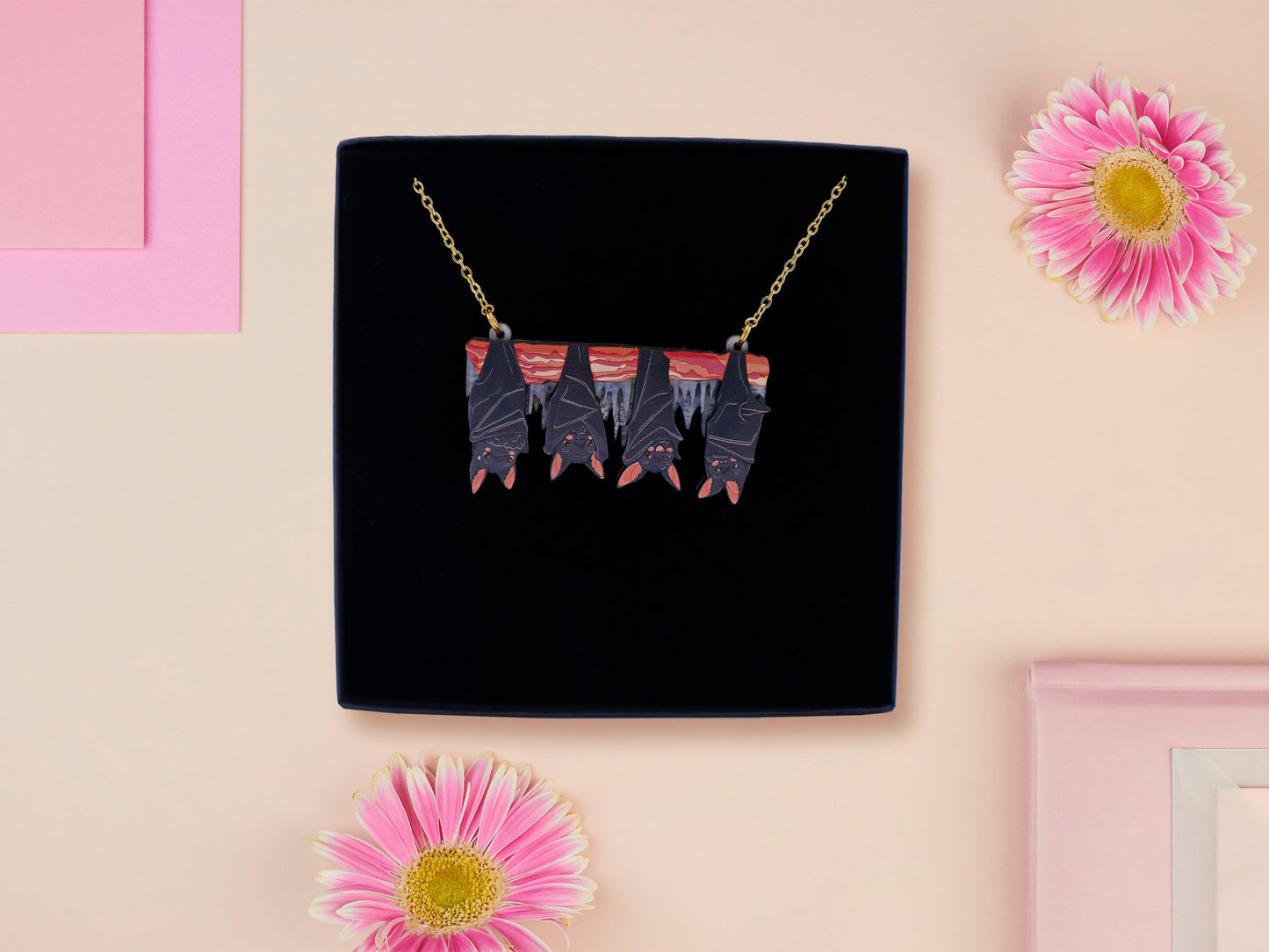 A wooden and glitter acrylic necklace with gold chain in a black box of four cute bats hung upside down on a cave wall of stalactites