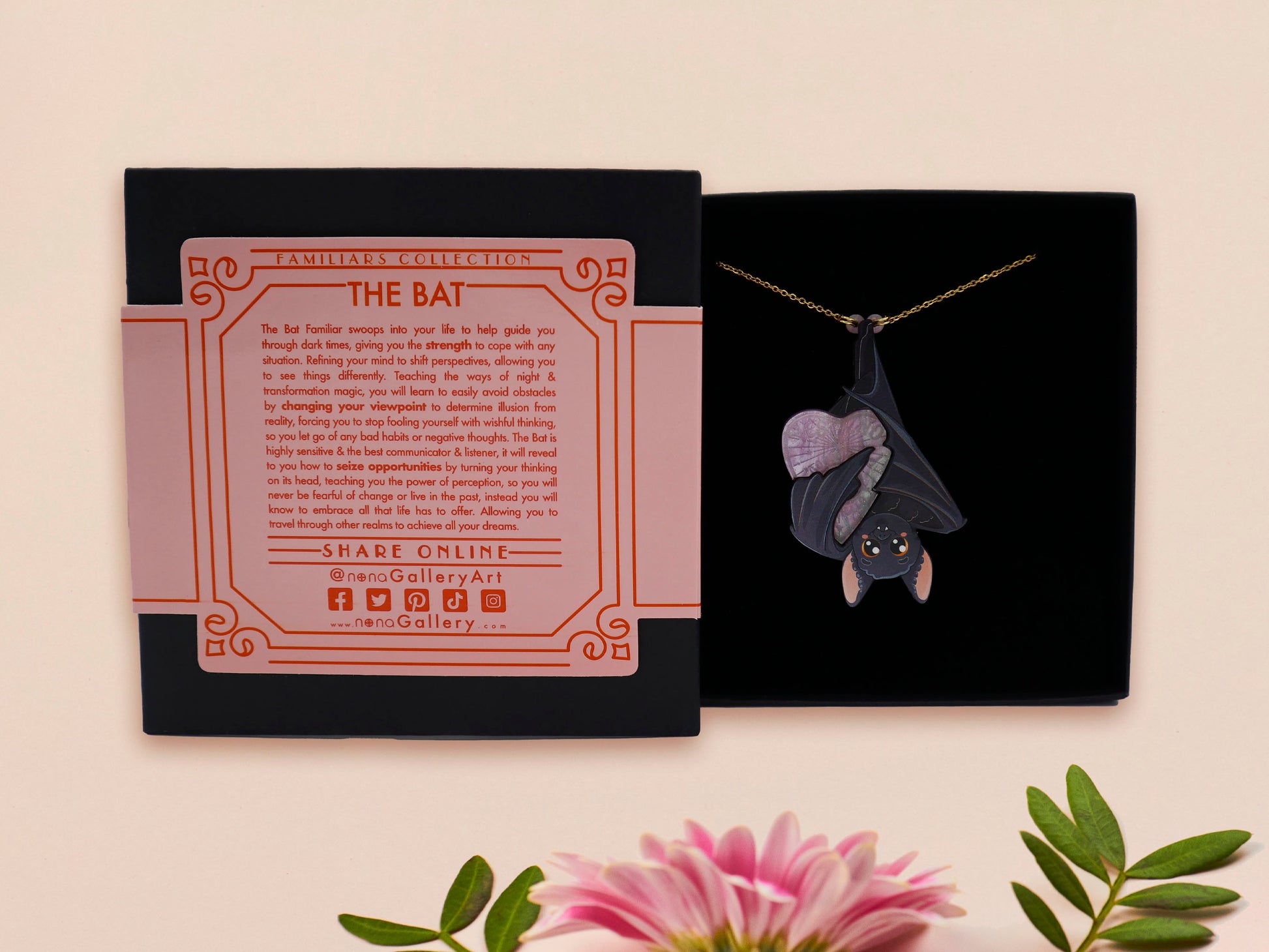 Mixed material handmade necklace of chibi cartoon bat hanging upside down holding a pearlescent Ouija planchette, with a gold chain and black gift box with a pink familiars collection gift sleeve.