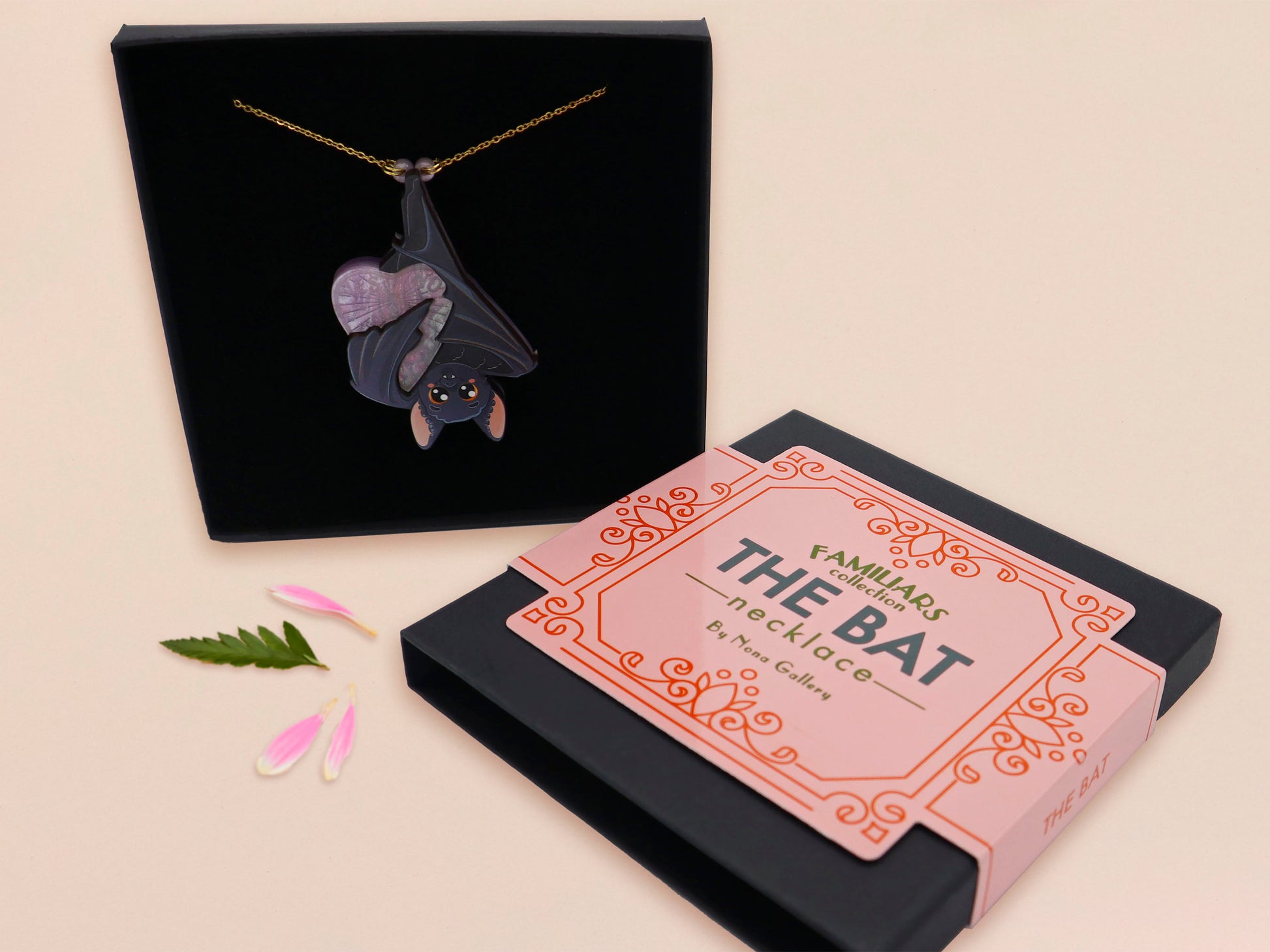 Mixed material handmade necklace of chibi cartoon bat hanging upside down holding a pearlescent Ouija planchette, with a gold chain and black gift box with a pink familiars collection gift sleeve.