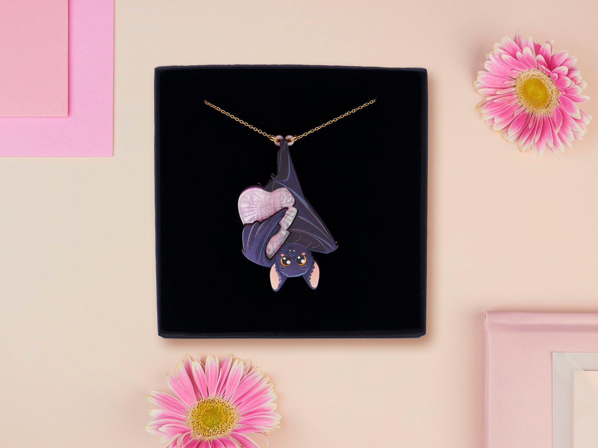 Mixed material handmade necklace of chibi cartoon bat hanging upside down holding a pearlescent Ouija planchette, with a gold chain and black gift box.