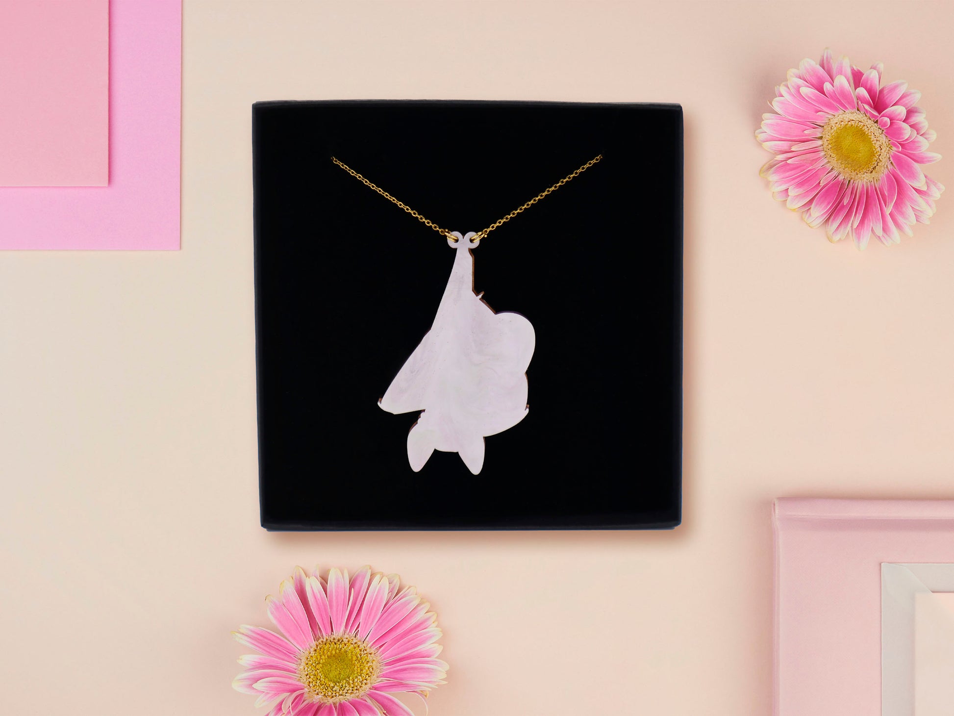 Back of mixed material handmade necklace of chibi cartoon bat hanging upside down holding a pearlescent Ouija planchette, with a gold chain and black gift box.