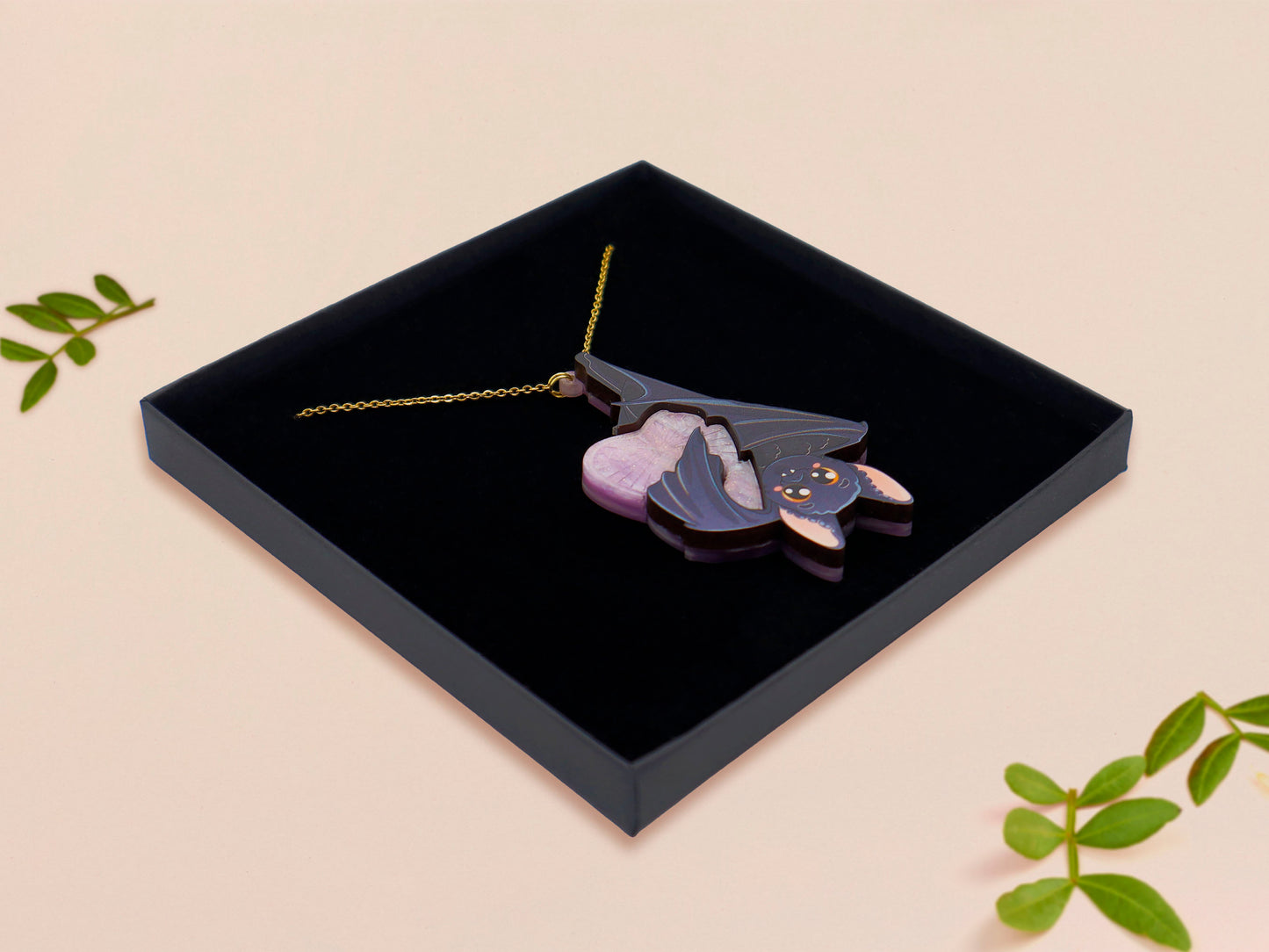 Mixed material handmade necklace of chibi cartoon bat hanging upside down holding a pearlescent Ouija planchette, with a gold chain and black gift box.