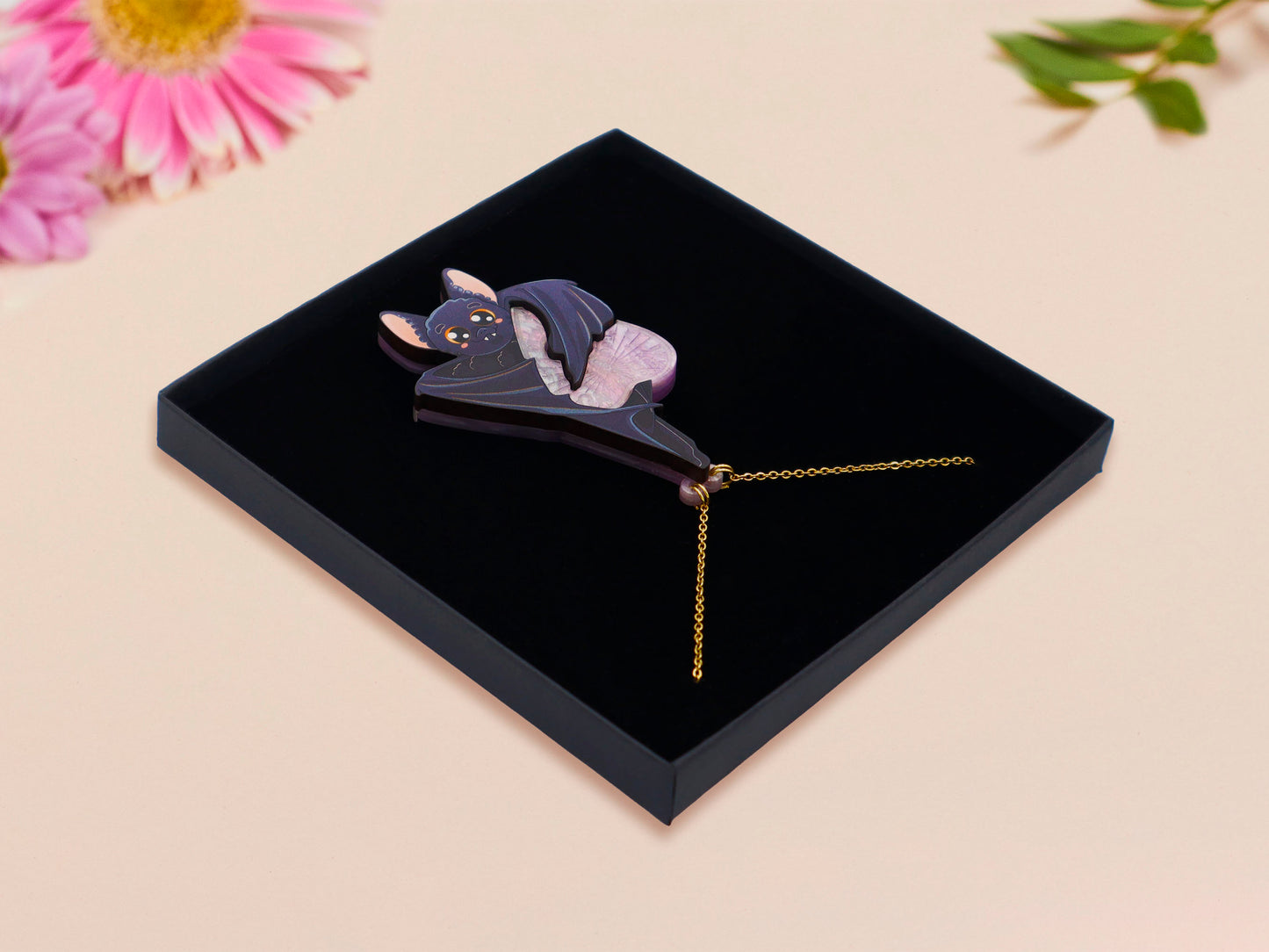 Mixed material handmade necklace of chibi cartoon bat hanging upside down holding a pearlescent Ouija planchette, with a gold chain and black gift box.