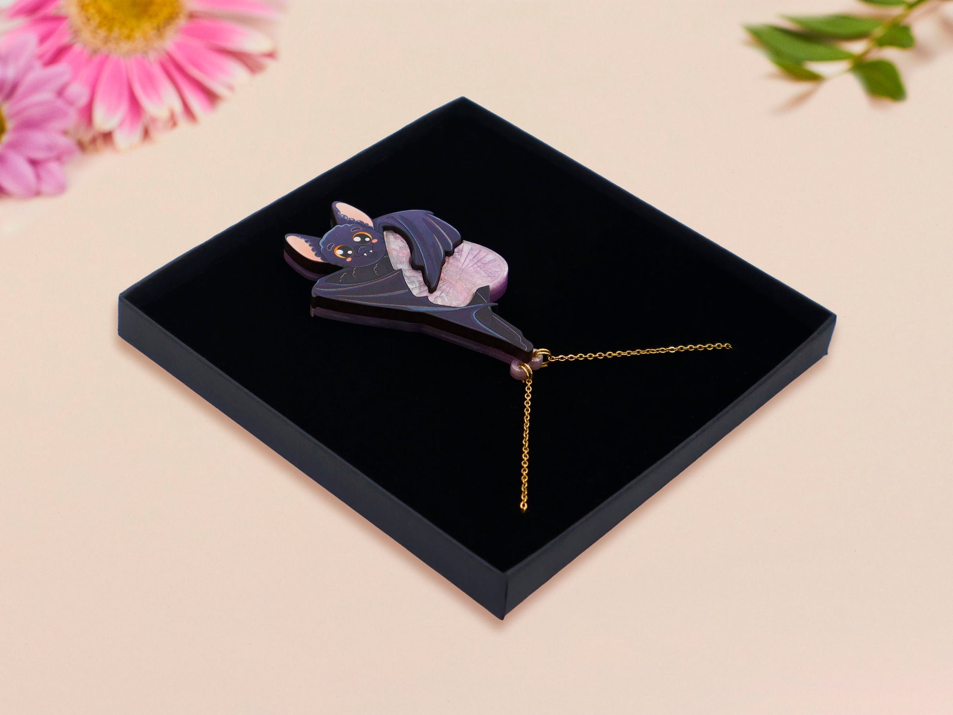 Mixed material handmade necklace of chibi cartoon bat hanging upside down holding a pearlescent Ouija planchette, with a gold chain and black gift box.