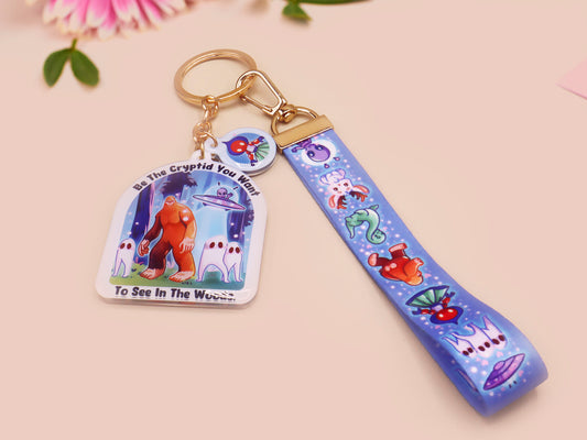 Acrylic epoxy cryptid and alien keychain with a large acrylic charm of a bigfoot yeti nightcrawlers and an alien in a UFO with the quote Be The Cryptidy You Want To See In the Woods, a smaller charm of the flatwood monster and a matching lanyard.