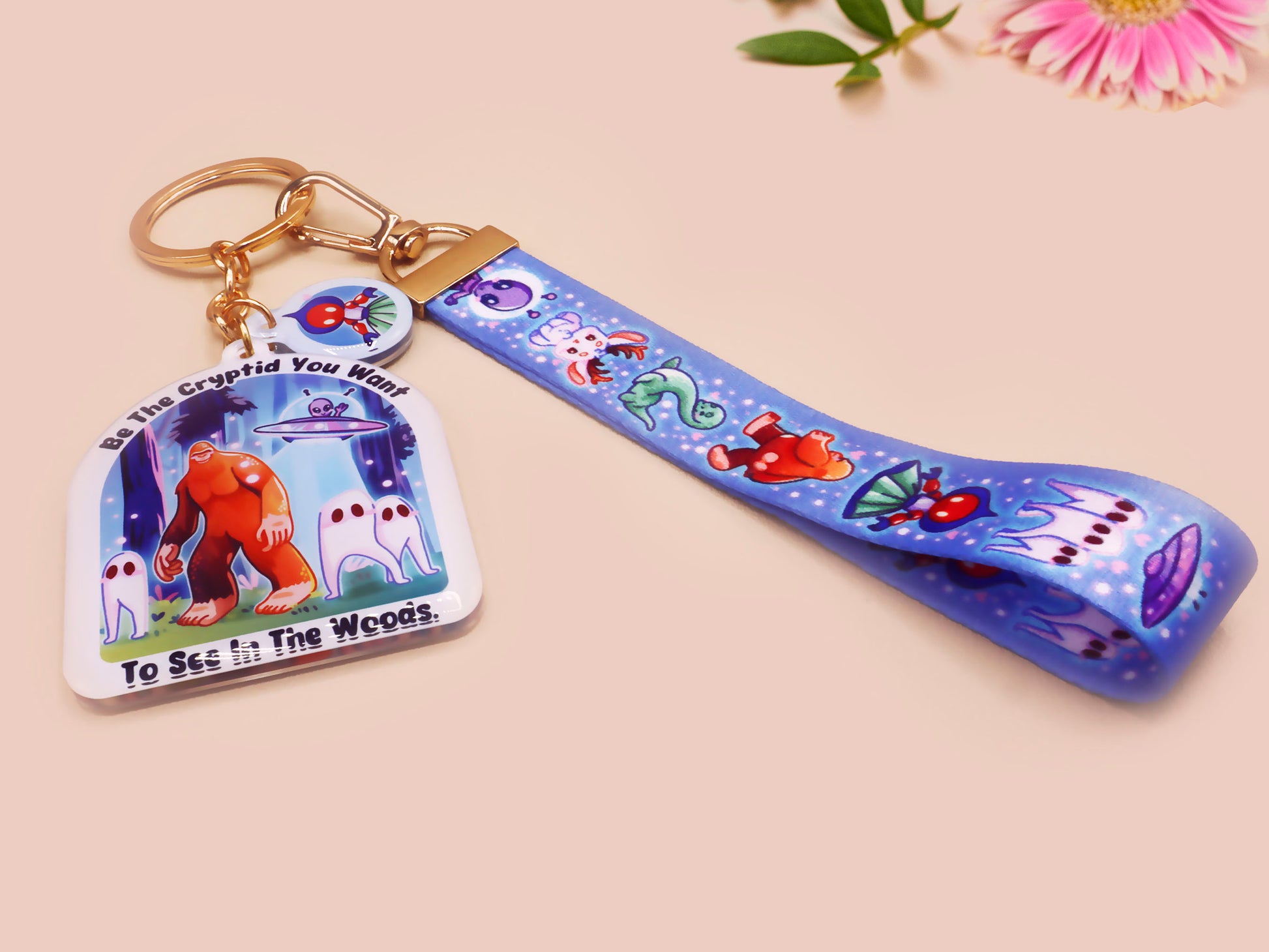 Acrylic epoxy cryptid and alien keychain with a large acrylic charm of a bigfoot yeti nightcrawlers and an alien in a UFO with the quote Be The Cryptidy You Want To See In the Woods, a smaller charm of the flatwood monster and a matching lanyard.