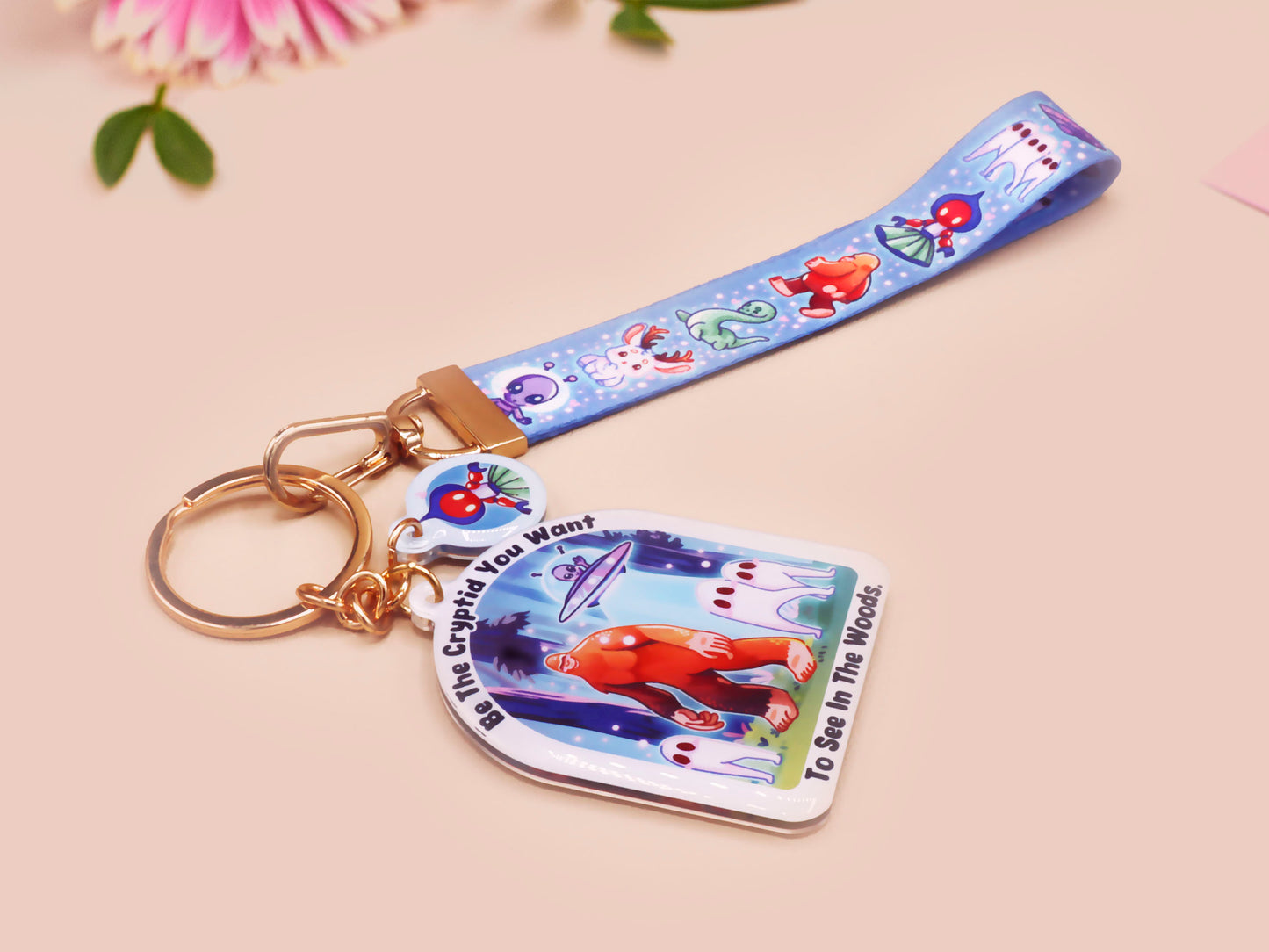 Acrylic epoxy cryptid and alien keychain with a large acrylic charm of a bigfoot yeti nightcrawlers and an alien in a UFO with the quote Be The Cryptidy You Want To See In the Woods, a smaller charm of the flatwood monster and a matching lanyard.