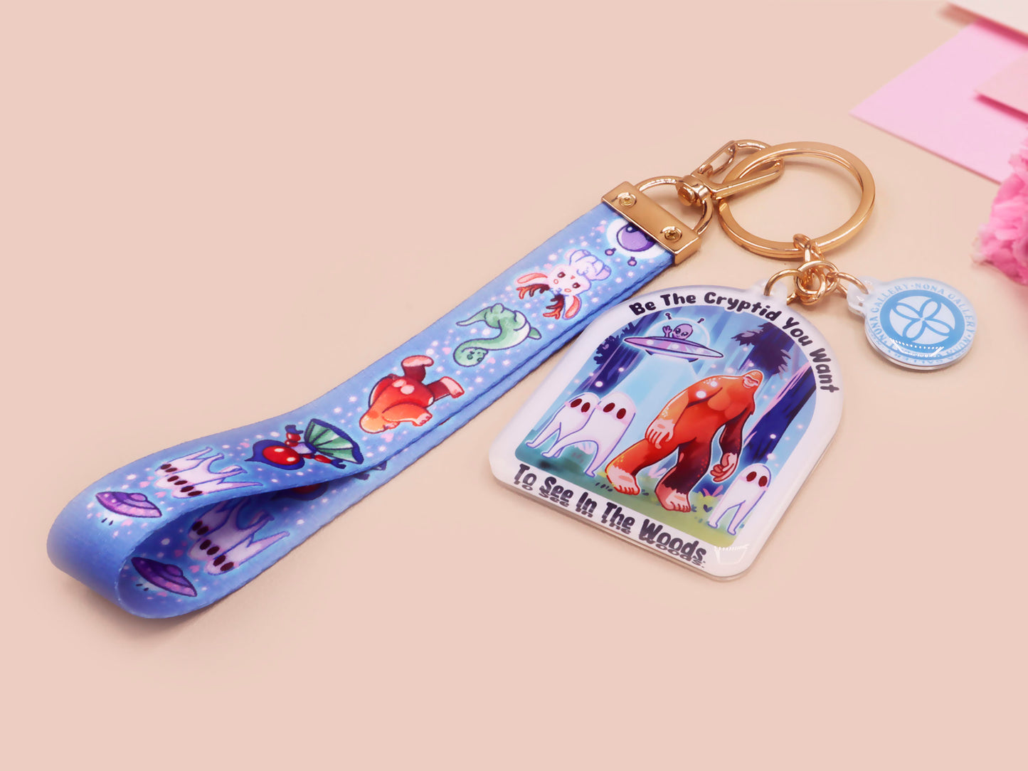 Acrylic epoxy cryptid and alien keychain with a large acrylic charm of a bigfoot yeti nightcrawlers and an alien in a UFO with the quote Be The Cryptidy You Want To See In the Woods, a smaller charm of the flatwood monster and a matching lanyard.