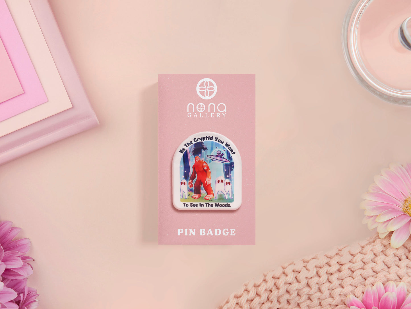 Clear acrylic pin badge with pink heart-shaped rubber pin back clasp. The design of the pin badge is a cute cartoon chibi fresno nightcrawlers, yeti bigfoot and alien UFO in a forest with the quote Be The Cryptid You Want To See In The Woods.