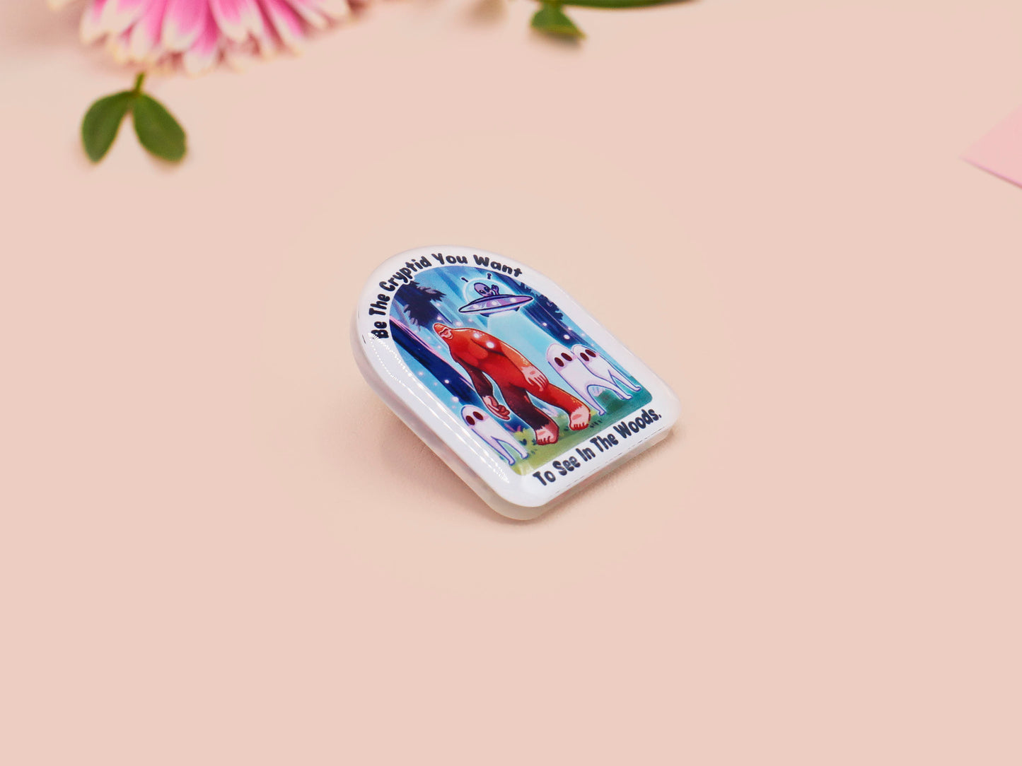 Clear acrylic pin badge with pink heart-shaped rubber pin back clasp. The design of the pin badge is a cute cartoon chibi fresno nightcrawlers, yeti bigfoot and alien UFO in a forest with the quote Be The Cryptid You Want To See In The Woods.