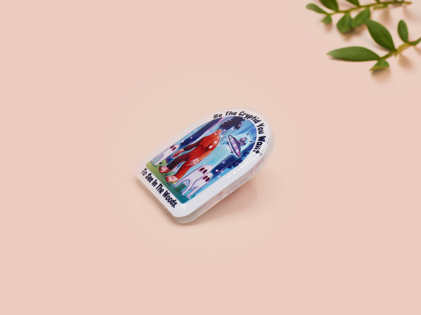 Clear acrylic pin badge with pink heart-shaped rubber pin back clasp. The design of the pin badge is a cute cartoon chibi fresno nightcrawlers, yeti bigfoot and alien UFO in a forest with the quote Be The Cryptid You Want To See In The Woods.
