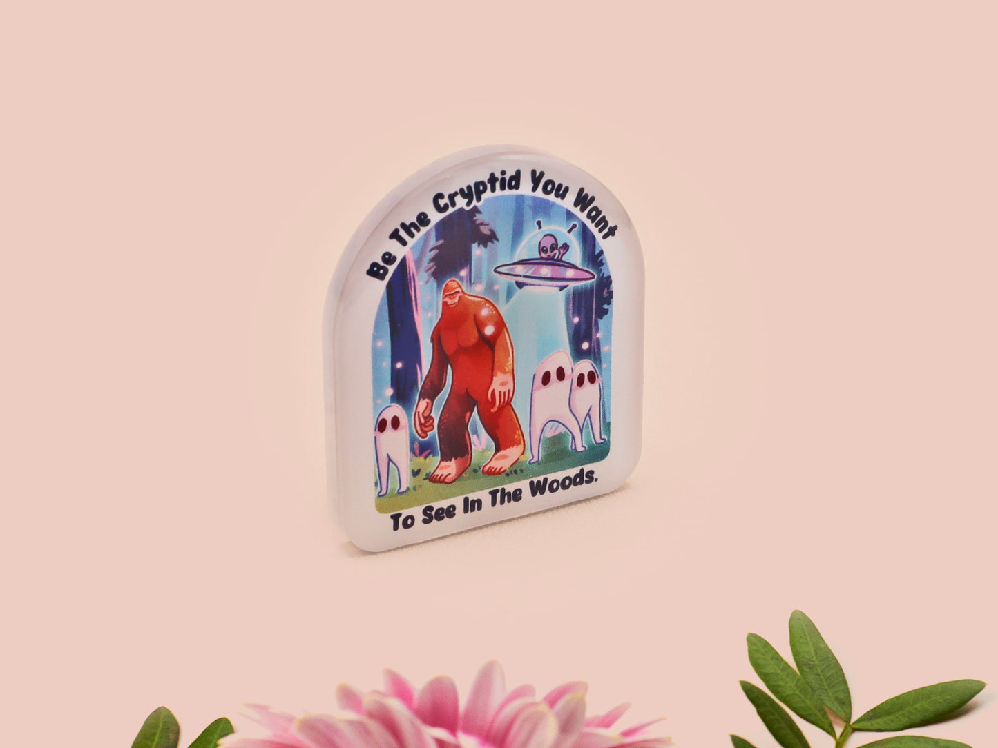 Clear acrylic pin badge with pink heart-shaped rubber pin back clasp. The design of the pin badge is a cute cartoon chibi fresno nightcrawlers, yeti bigfoot and alien UFO in a forest with the quote Be The Cryptid You Want To See In The Woods.