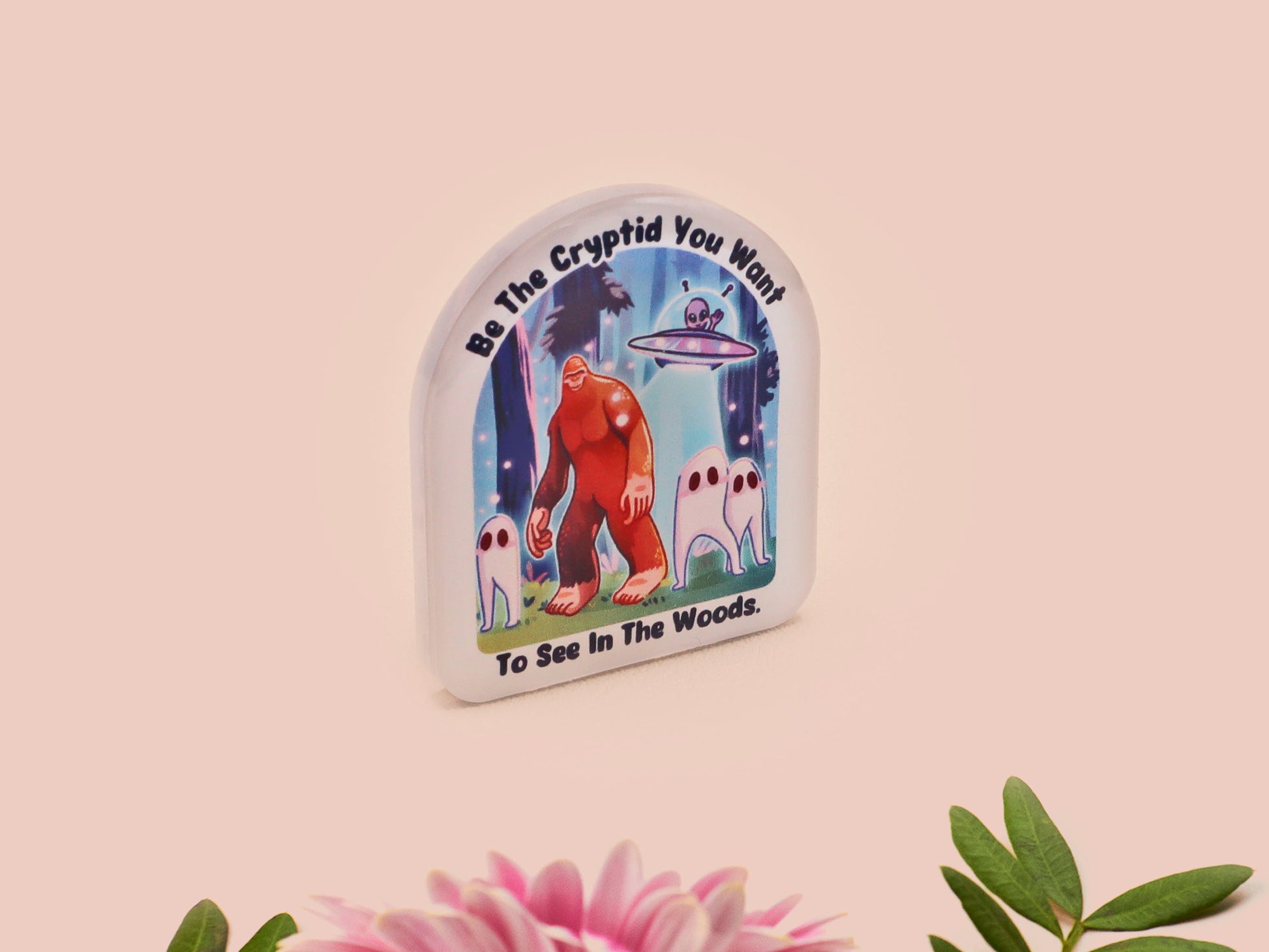Clear acrylic pin badge with pink heart-shaped rubber pin back clasp. The design of the pin badge is a cute cartoon chibi fresno nightcrawlers, yeti bigfoot and alien UFO in a forest with the quote Be The Cryptid You Want To See In The Woods.