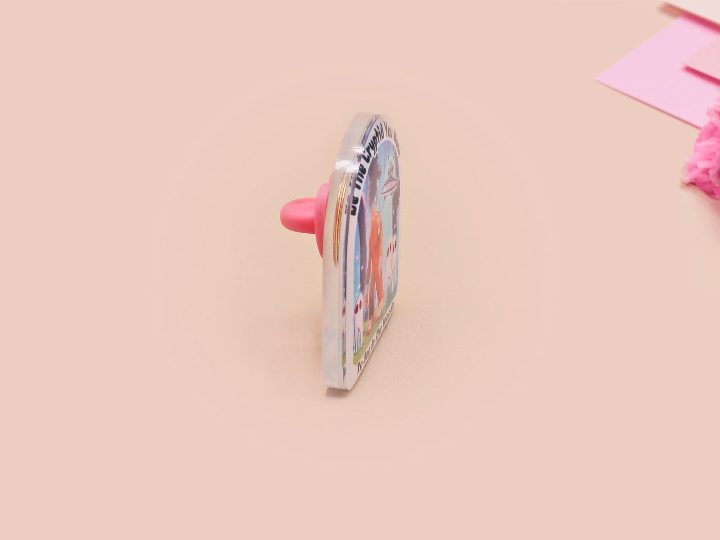 Clear acrylic pin badge with pink heart-shaped rubber pin back clasp. The design of the pin badge is a cute cartoon chibi fresno nightcrawlers, yeti bigfoot and alien UFO in a forest with the quote Be The Cryptid You Want To See In The Woods.