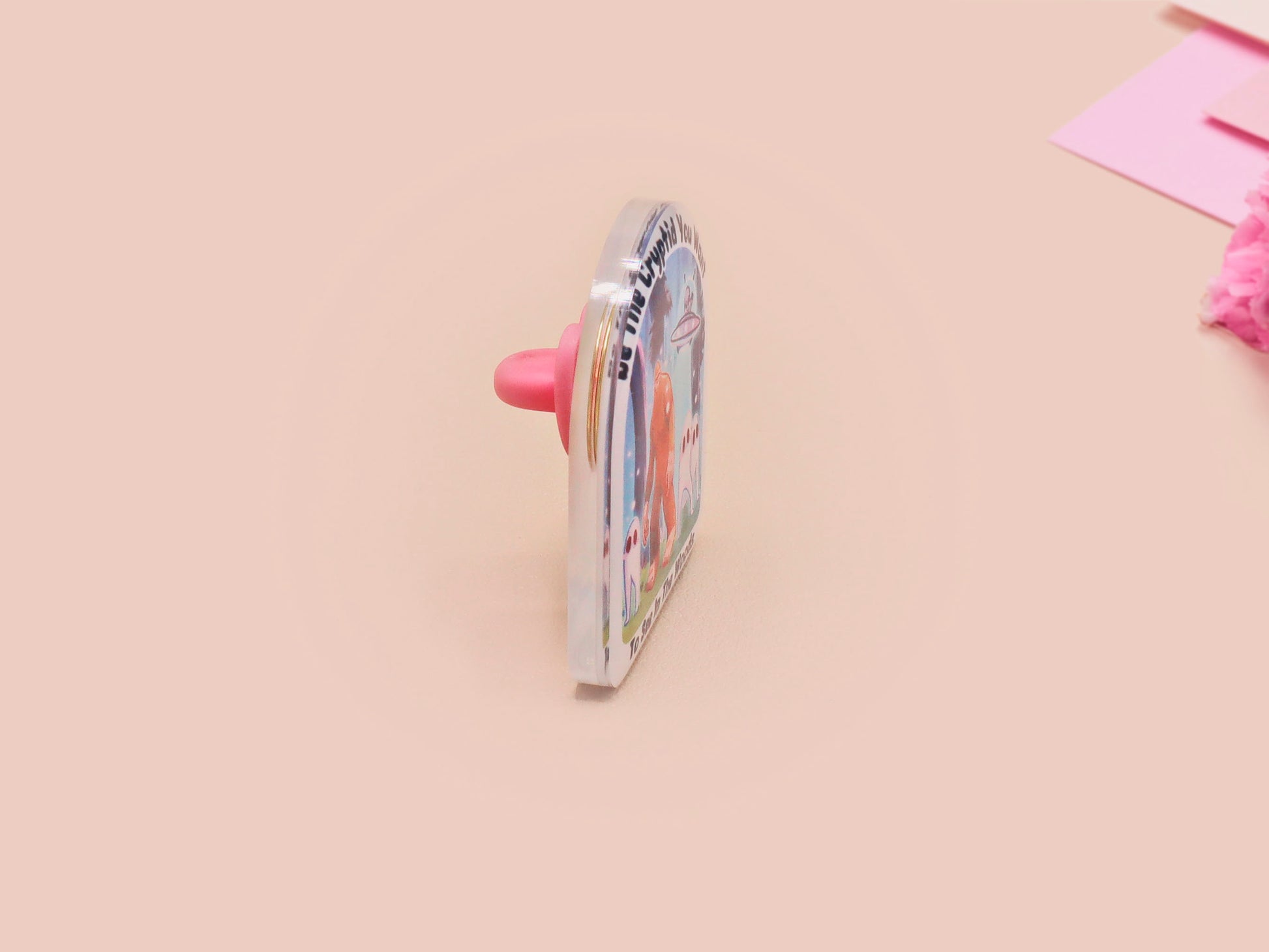 Clear acrylic pin badge with pink heart-shaped rubber pin back clasp. The design of the pin badge is a cute cartoon chibi fresno nightcrawlers, yeti bigfoot and alien UFO in a forest with the quote Be The Cryptid You Want To See In The Woods.