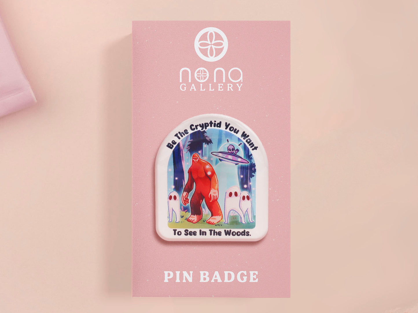 Clear acrylic pin badge with pink heart-shaped rubber pin back clasp. The design of the pin badge is a cute cartoon chibi fresno nightcrawlers, yeti bigfoot and alien UFO in a forest with the quote Be The Cryptid You Want To See In The Woods.