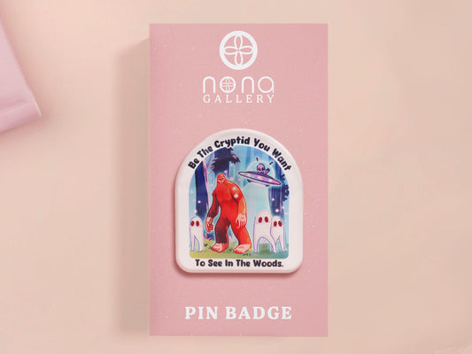 Clear acrylic pin badge with pink heart-shaped rubber pin back clasp. The design of the pin badge is a cute cartoon chibi fresno nightcrawlers, yeti bigfoot and alien UFO in a forest with the quote Be The Cryptid You Want To See In The Woods.