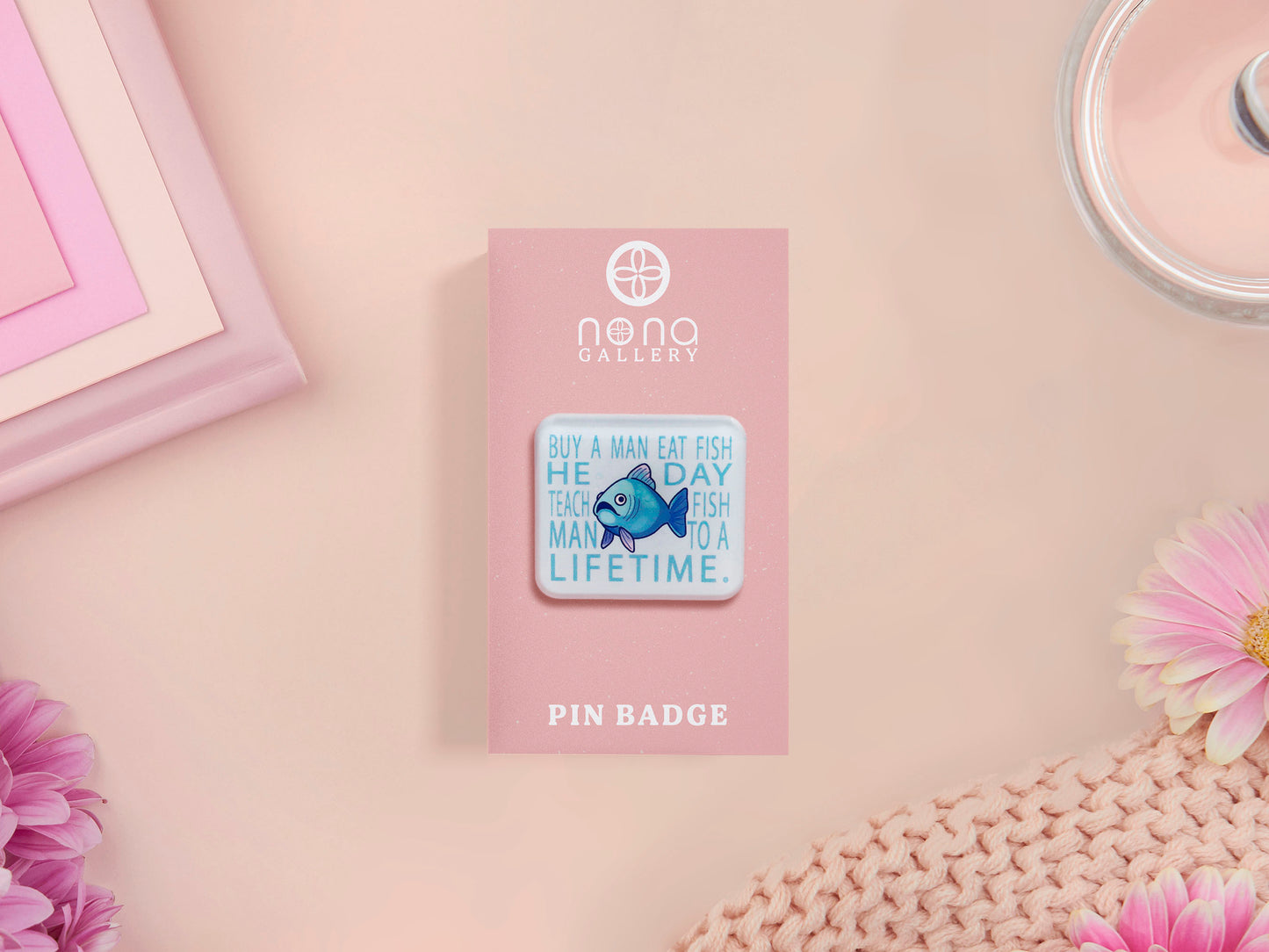 A clear acrylic pin badge with a pink heart-shaped pin back clasp. The design of the pin badge is of a cute chibi blue fish with the quote buy a man eat fish he day teach fish man to a lifetime.