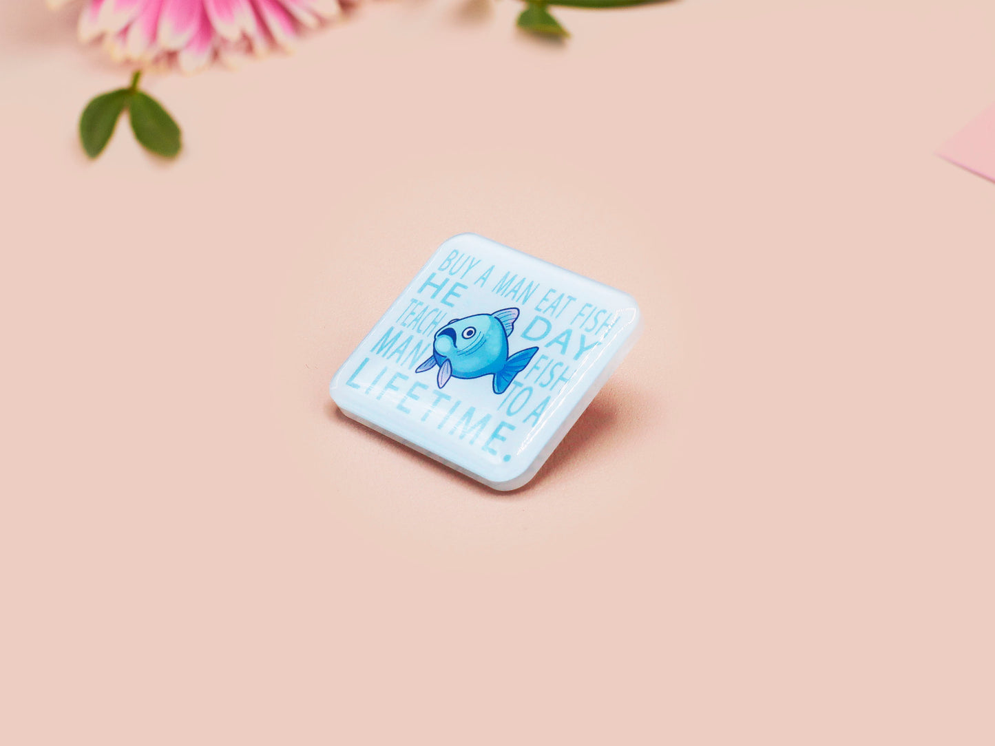 A clear acrylic pin badge with a pink heart-shaped pin back clasp. The design of the pin badge is of a cute chibi blue fish with the quote buy a man eat fish he day teach fish man to a lifetime.