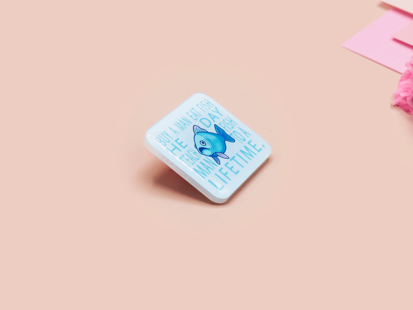 A clear acrylic pin badge with a pink heart-shaped pin back clasp. The design of the pin badge is of a cute chibi blue fish with the quote buy a man eat fish he day teach fish man to a lifetime.