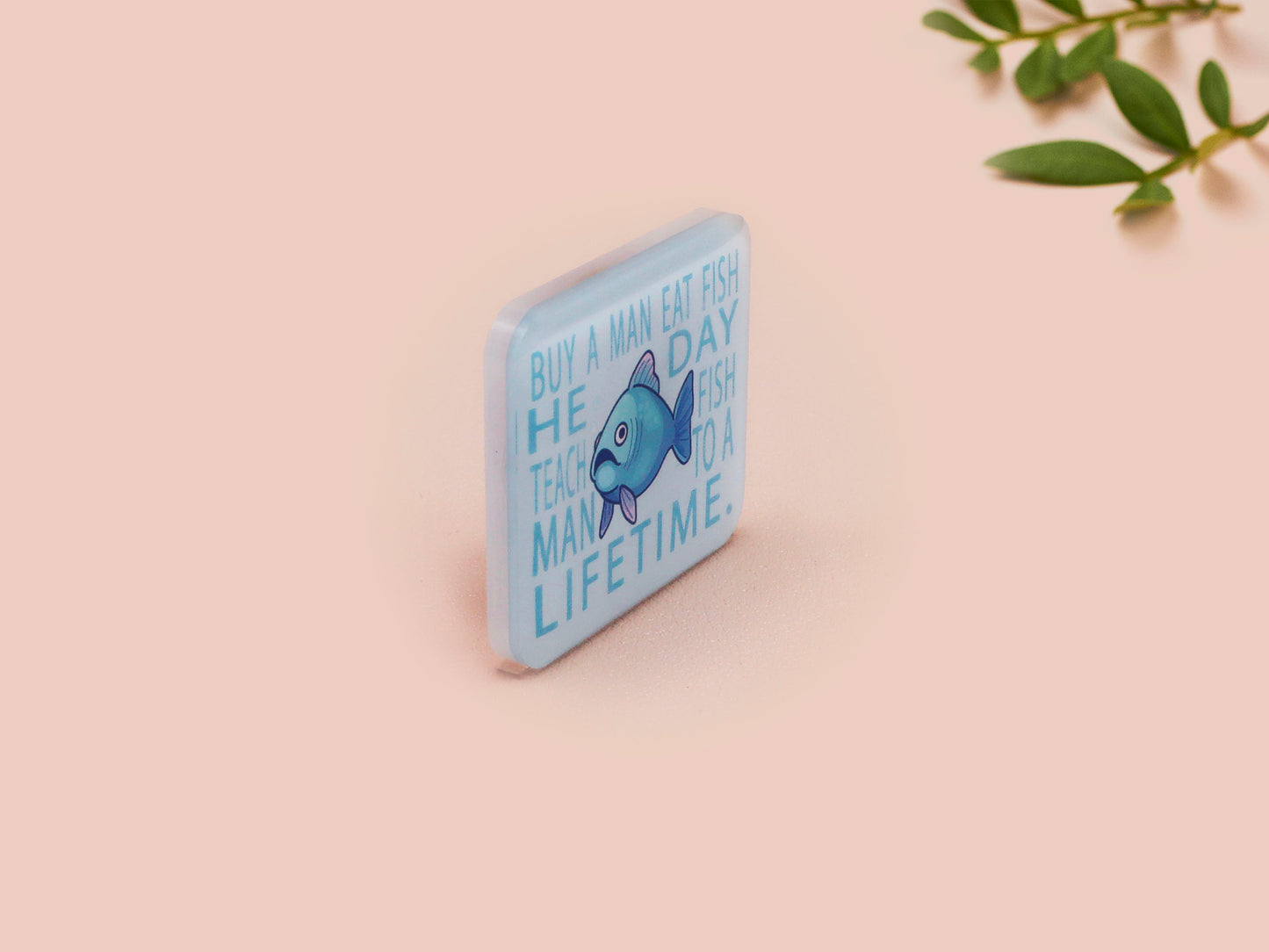 A clear acrylic pin badge with a pink heart-shaped pin back clasp. The design of the pin badge is of a cute chibi blue fish with the quote buy a man eat fish he day teach fish man to a lifetime.