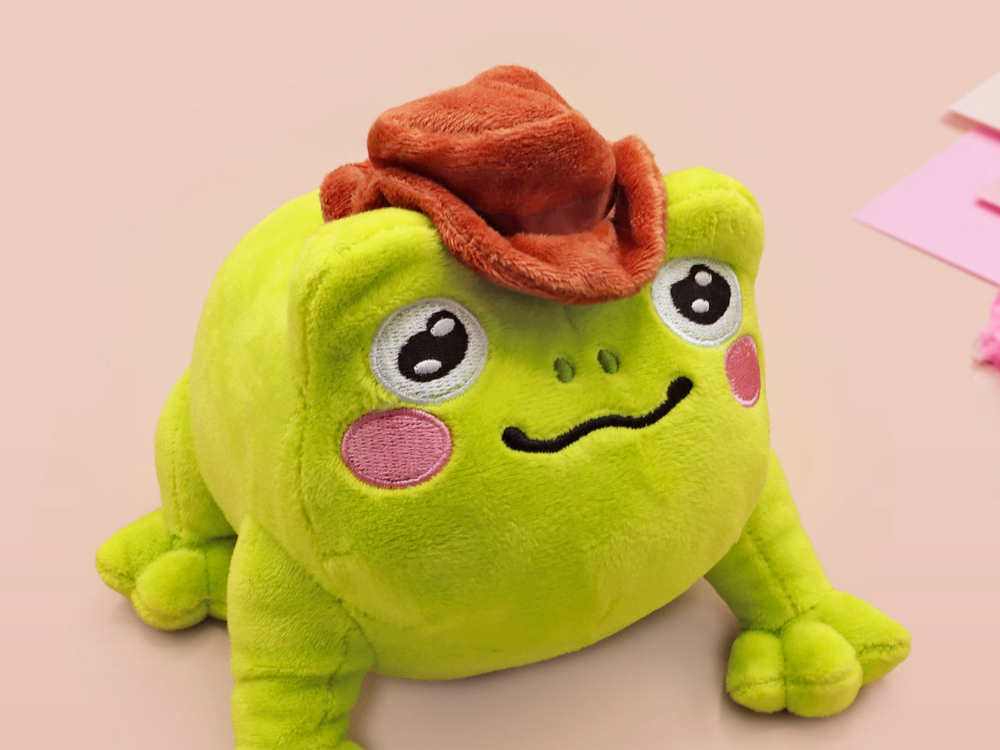 An adorable green frog plushie toy with cute embroidered eyes and facial expression, wearing a cowboy hat with a brown ribbon, and a hang tag describing the frog as Carl.
