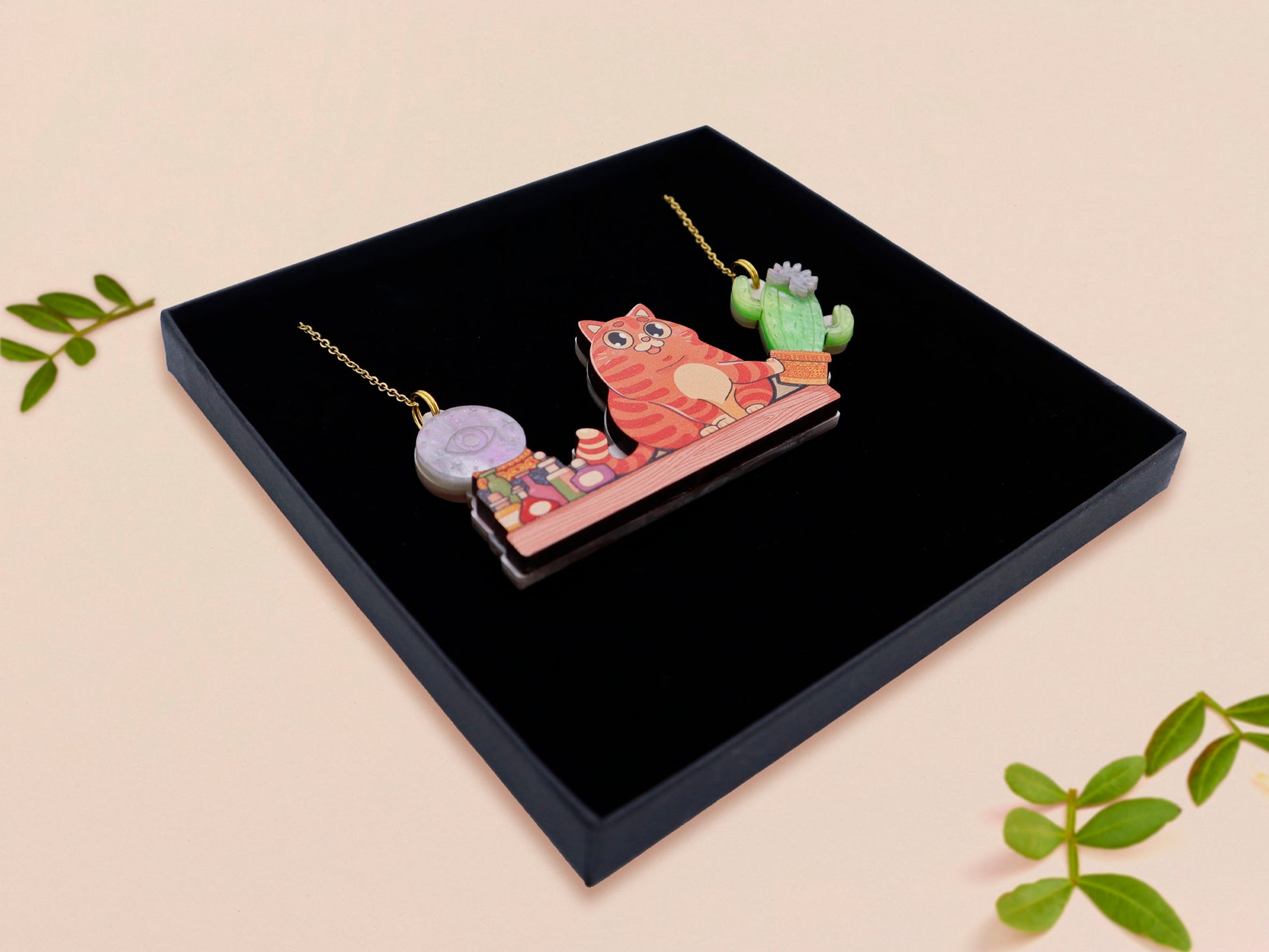 A wooden and glitter acrylic necklace with gold chain in a black box of a cute fat ginger cat sat on a shelf mischievously pushing a cactus of the end of the shelf, whilst sat next to a crystal ball and potions
