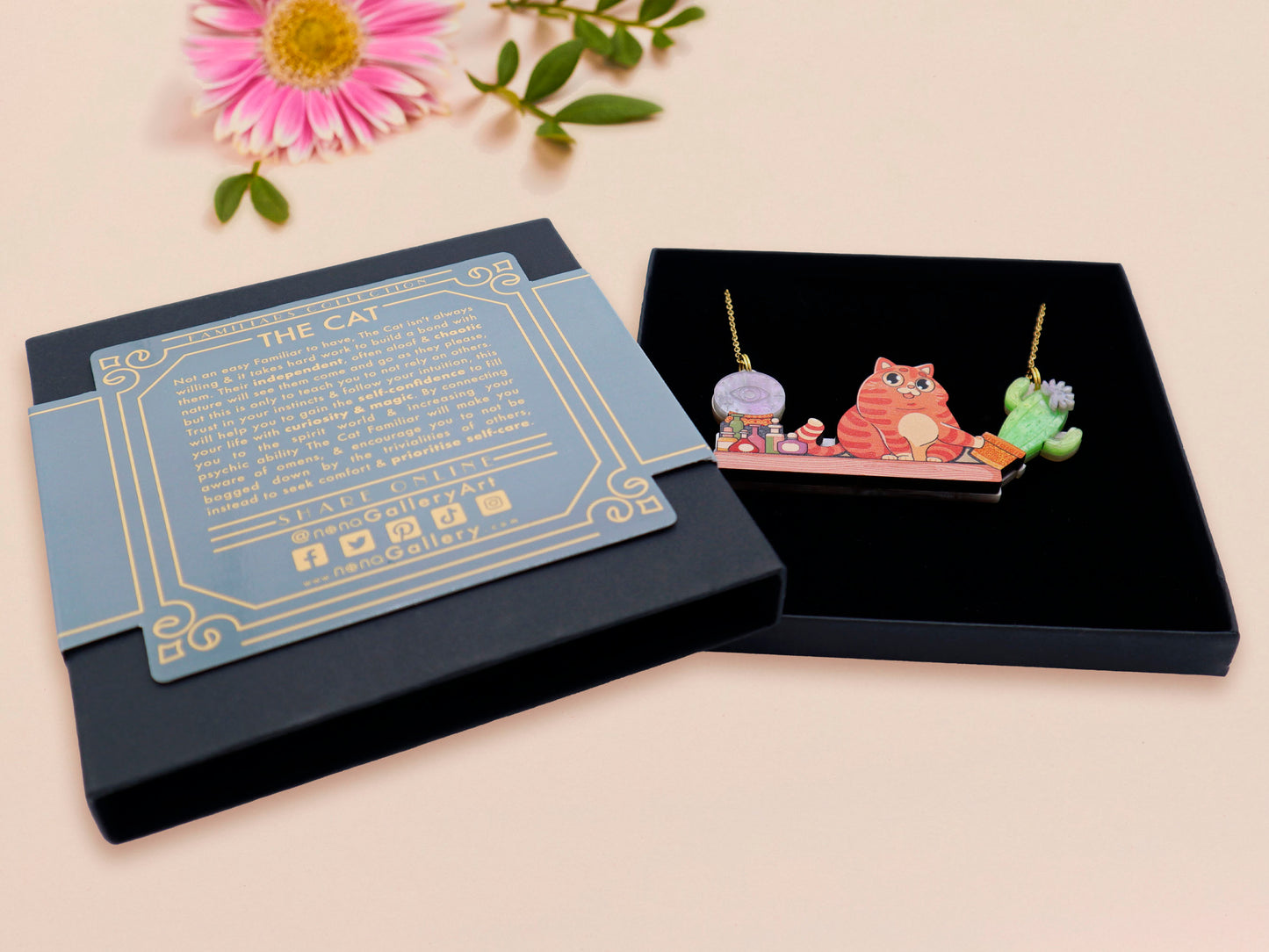 A wooden and glitter acrylic necklace with gold chain in a black box of a cute fat ginger cat sat on a shelf mischievously pushing a cactus of the end of the shelf, whilst sat next to a crystal ball and potions