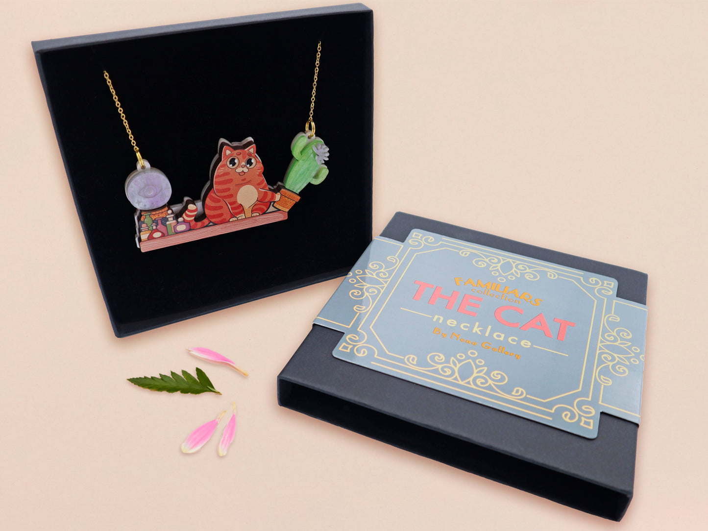 A wooden and glitter acrylic necklace with gold chain in a black box of a cute fat ginger cat sat on a shelf mischievously pushing a cactus of the end of the shelf, whilst sat next to a crystal ball and potions