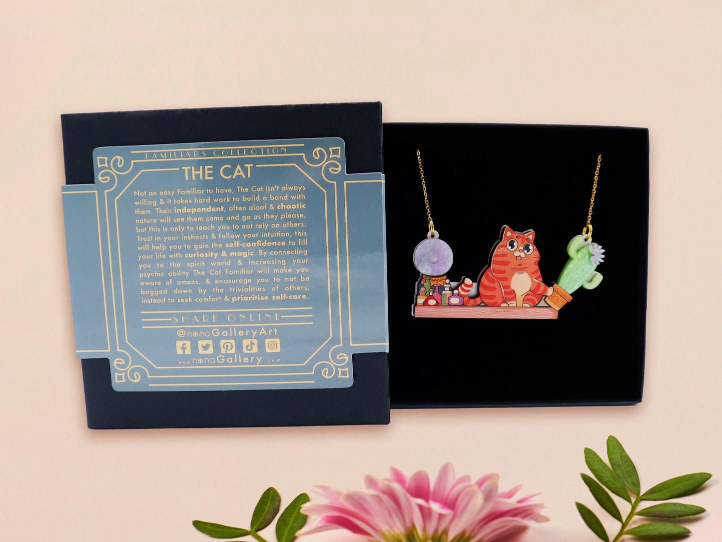A wooden and glitter acrylic necklace with gold chain in a black box of a cute fat ginger cat sat on a shelf mischievously pushing a cactus of the end of the shelf, whilst sat next to a crystal ball and potions