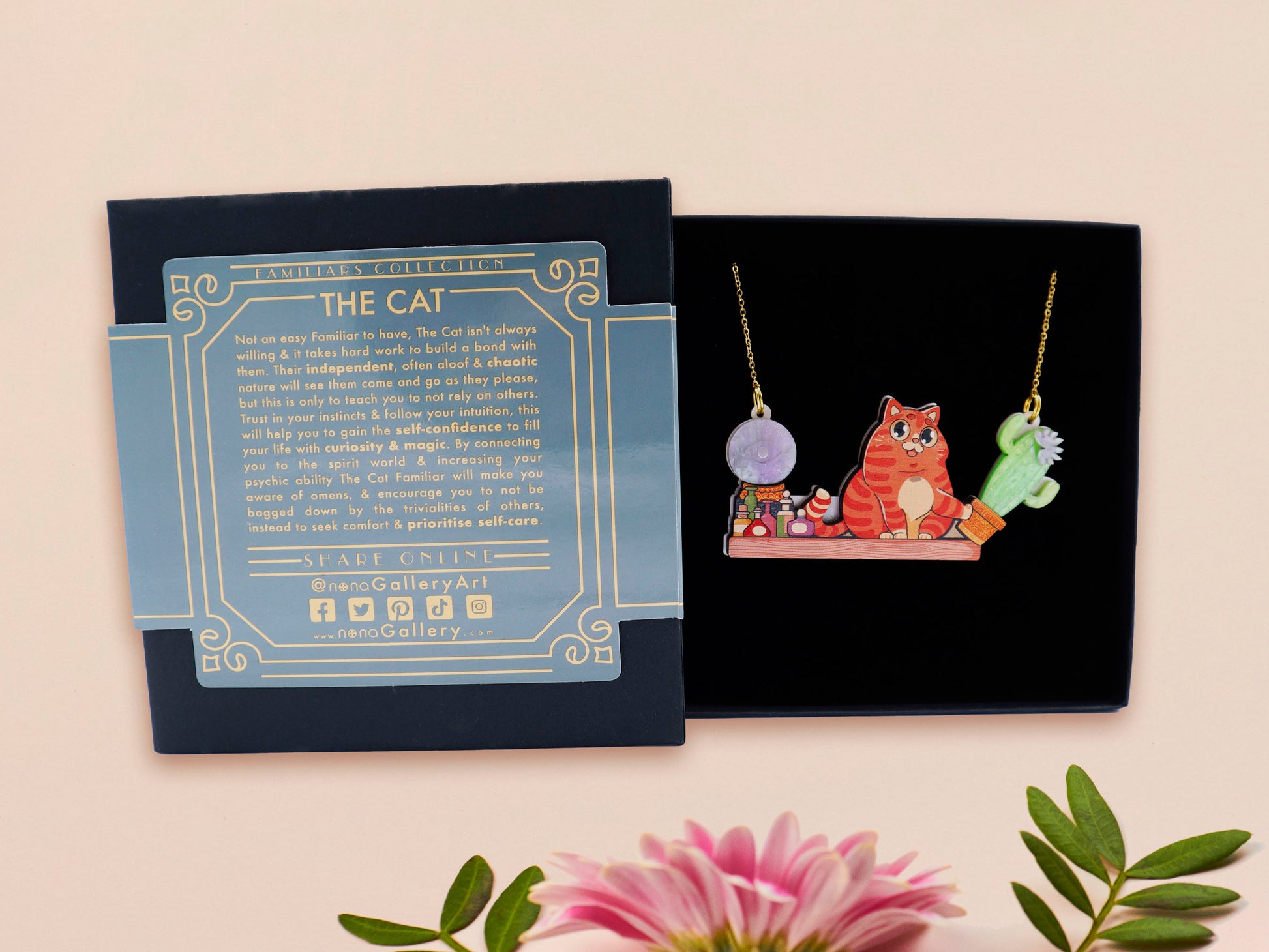 A wooden and glitter acrylic necklace with gold chain in a black box of a cute fat ginger cat sat on a shelf mischievously pushing a cactus of the end of the shelf, whilst sat next to a crystal ball and potions