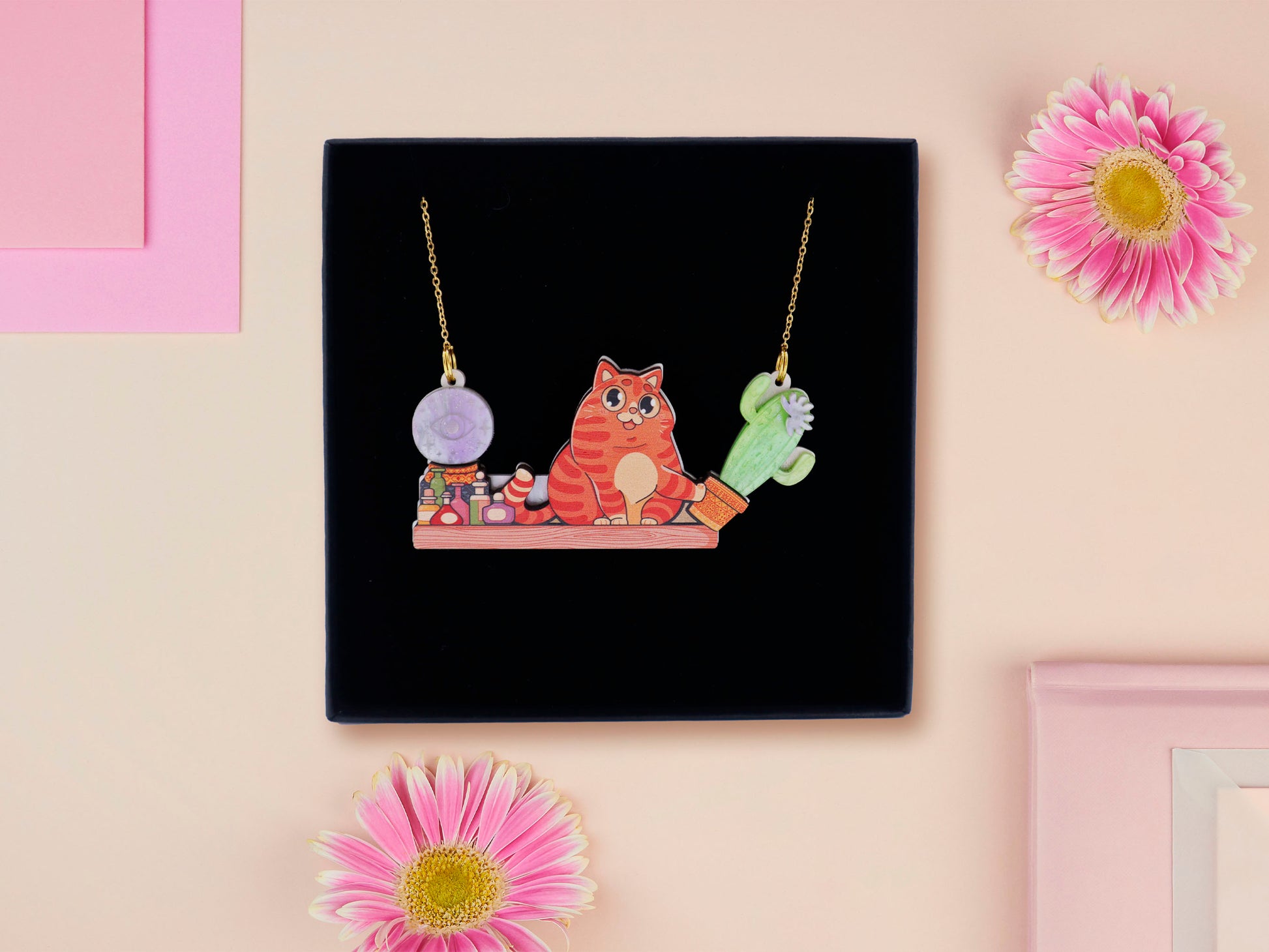 A wooden and glitter acrylic necklace with gold chain in a black box of a cute fat ginger cat sat on a shelf mischievously pushing a cactus of the end of the shelf, whilst sat next to a crystal ball and potions