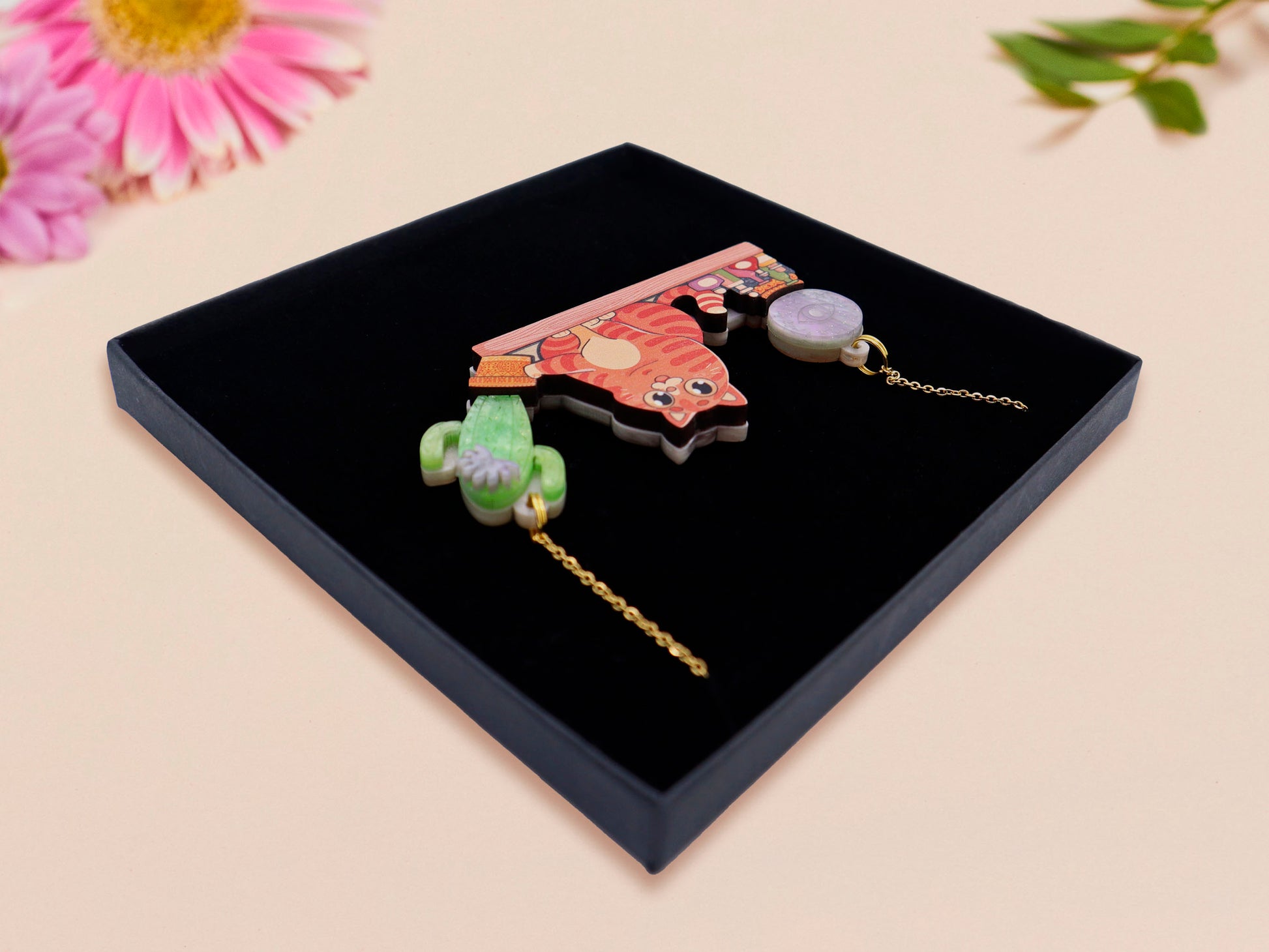 A wooden and glitter acrylic necklace with gold chain in a black box of a cute fat ginger cat sat on a shelf mischievously pushing a cactus of the end of the shelf, whilst sat next to a crystal ball and potions