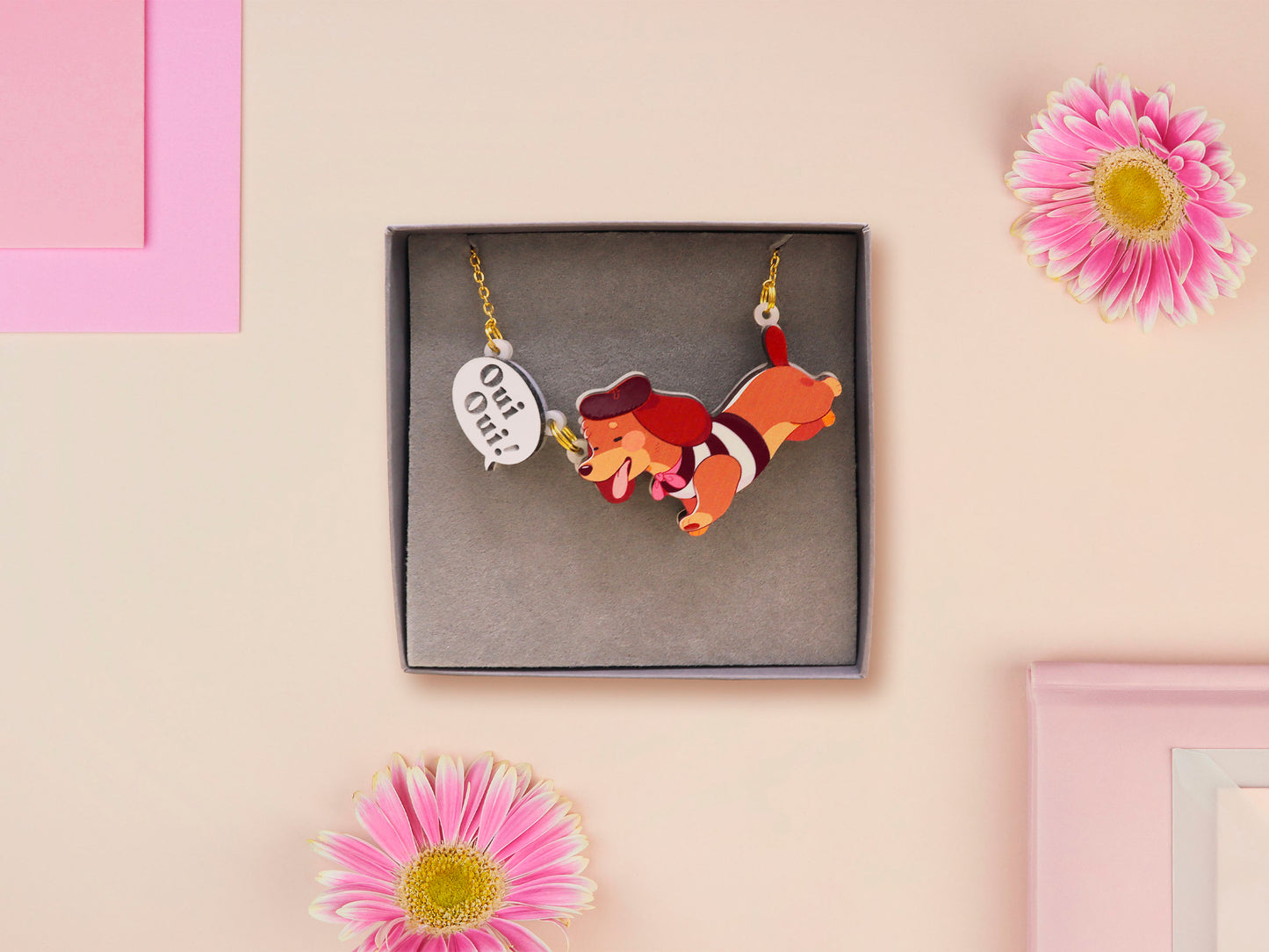 An adorable cute dachshund sausage dog necklace in a french outfit, a beret and striped top, connected to a speech bubble that is engraved with the words oui oui! displayed in a velvet gift box with a unique cute dachshund sleeve design.