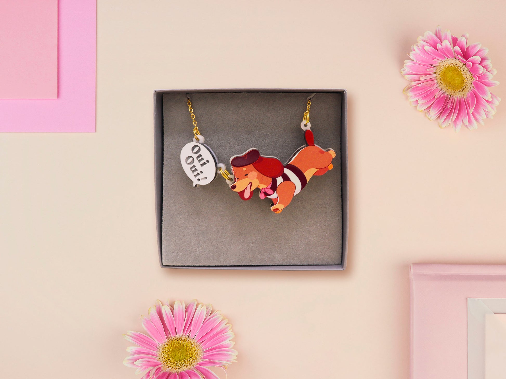 An adorable cute dachshund sausage dog necklace in a french outfit, a beret and striped top, connected to a speech bubble that is engraved with the words oui oui! displayed in a velvet gift box with a unique cute dachshund sleeve design.
