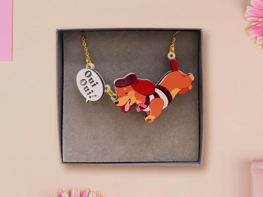 An adorable cute dachshund sausage dog necklace in a french outfit, a beret and a striped top, connected to a speech bubble that is engraved with the words oui oui! displayed in a velvet gift box with a unique cute dachshund sleeve design.