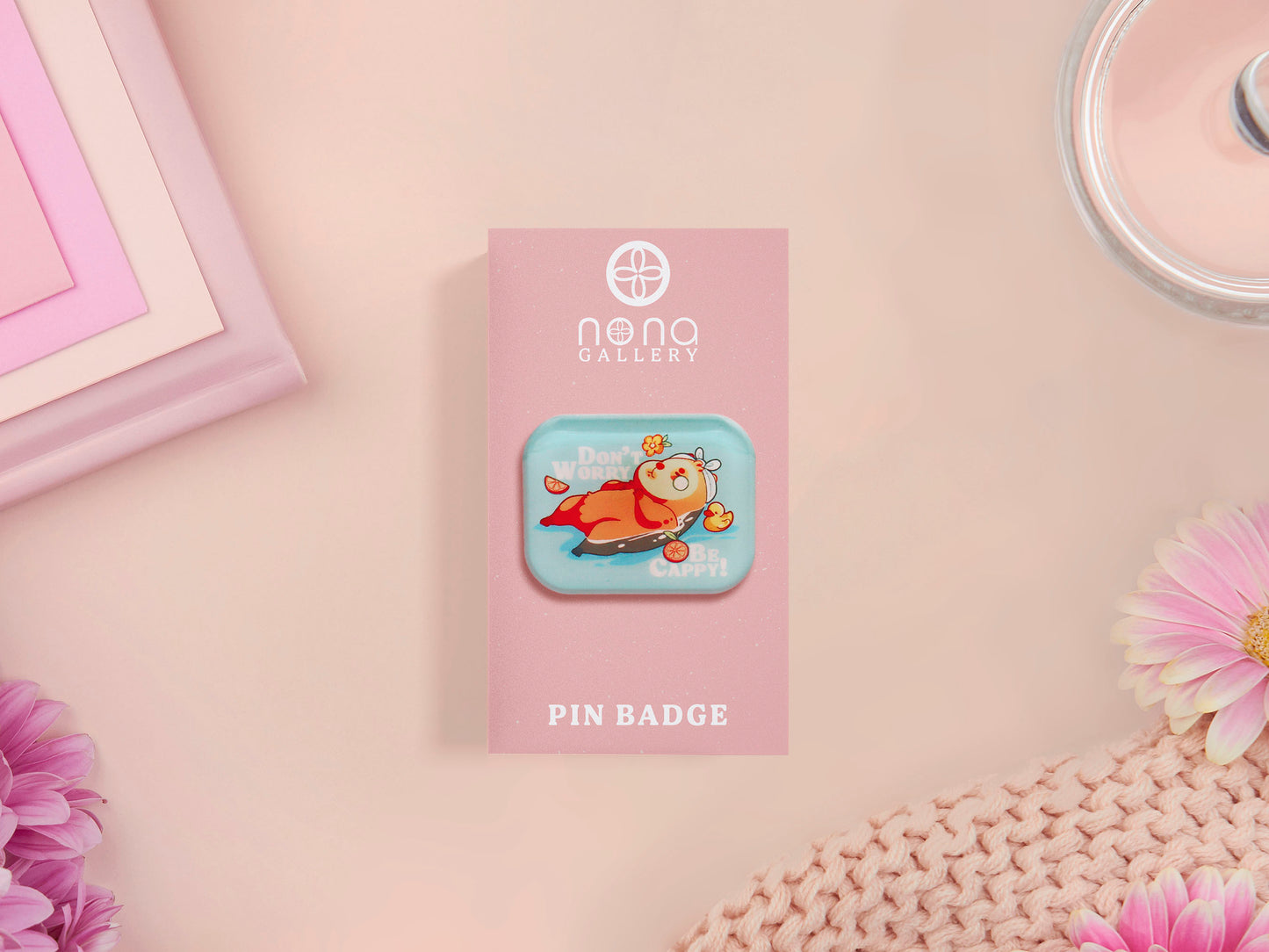 A clear acrylic pin badge with a pink heart-shaped pin back clasp. The design of the pin badge is of a cute relaxing capybara in water with a face mask with the quote don't worry be cappy.