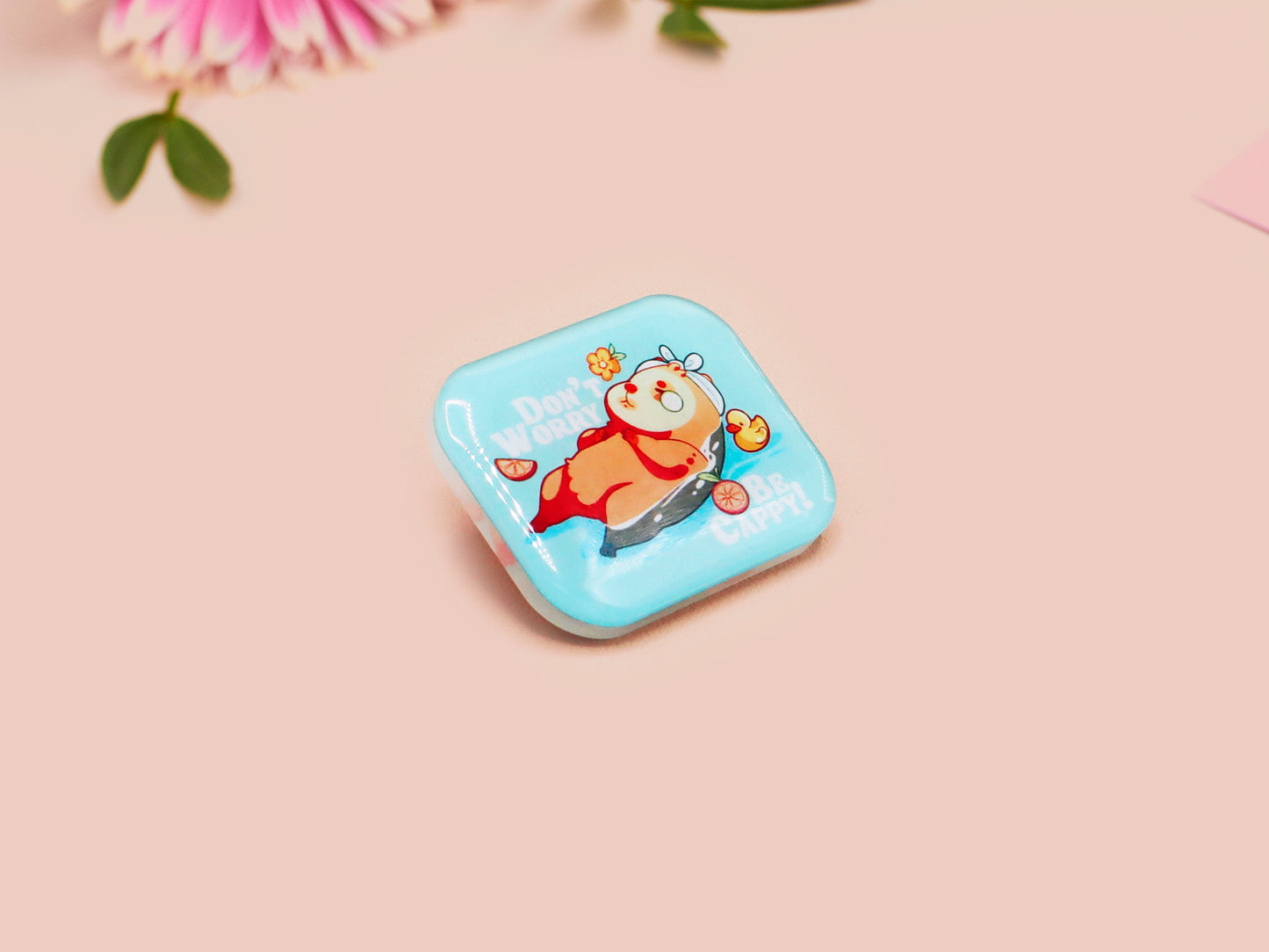 A clear acrylic pin badge with a pink heart-shaped pin back clasp. The design of the pin badge is of a cute relaxing capybara in water with a face mask with the quote don't worry be cappy.
