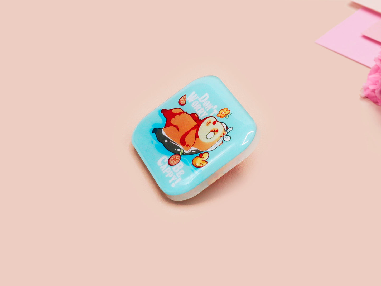 A clear acrylic pin badge with a pink heart-shaped pin back clasp. The design of the pin badge is of a cute relaxing capybara in water with a face mask with the quote don't worry be cappy.