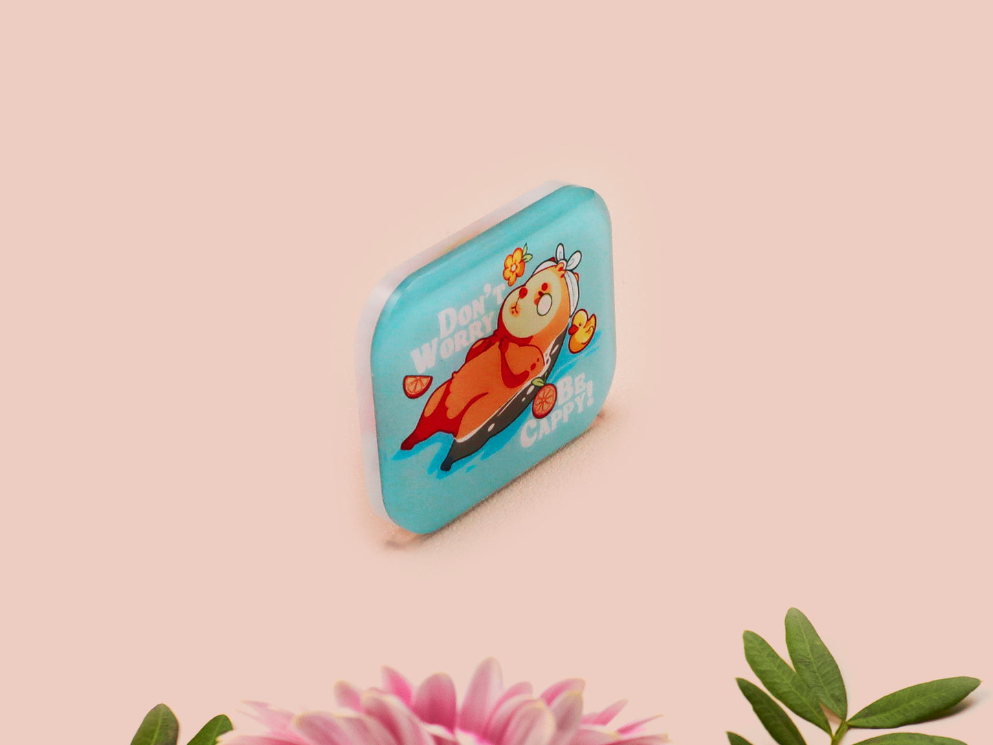 A clear acrylic pin badge with a pink heart-shaped pin back clasp. The design of the pin badge is of a cute relaxing capybara in water with a face mask with the quote don't worry be cappy.
