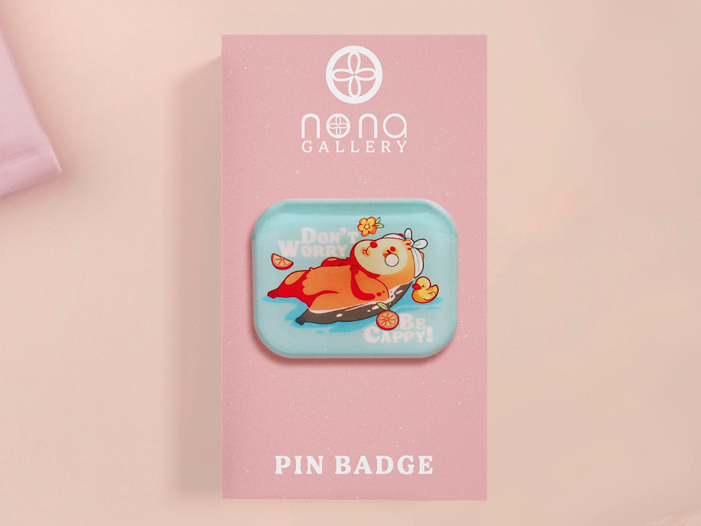 A clear acrylic pin badge with a pink heart-shaped pin back clasp. The design of the pin badge is of a cute relaxing capybara in water with a face mask with the quote don't worry be cappy.