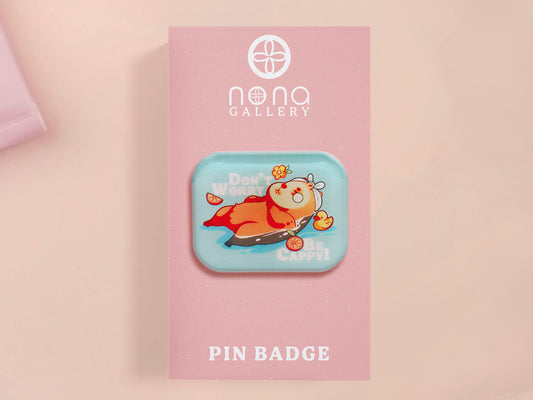 A clear acrylic pin badge with a pink heart-shaped pin back clasp. The design of the pin badge is of a cute relaxing capybara in water with a face mask with the quote don't worry be cappy.