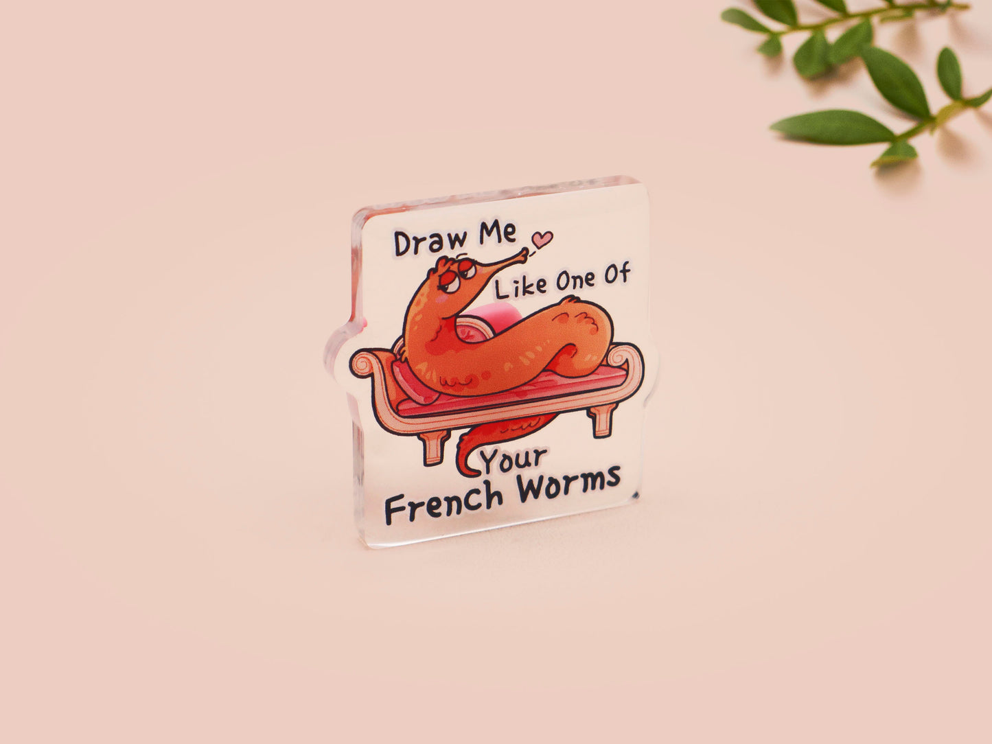 A cute transparent acrylic pin badge with a pink heart-shaped rubber clasp pin back. The design of the pin badge is a cartoon orange worm on a sting sitting on a sofa chaise lounge with the titanic quote Draw Me Like One of Your French Worms
