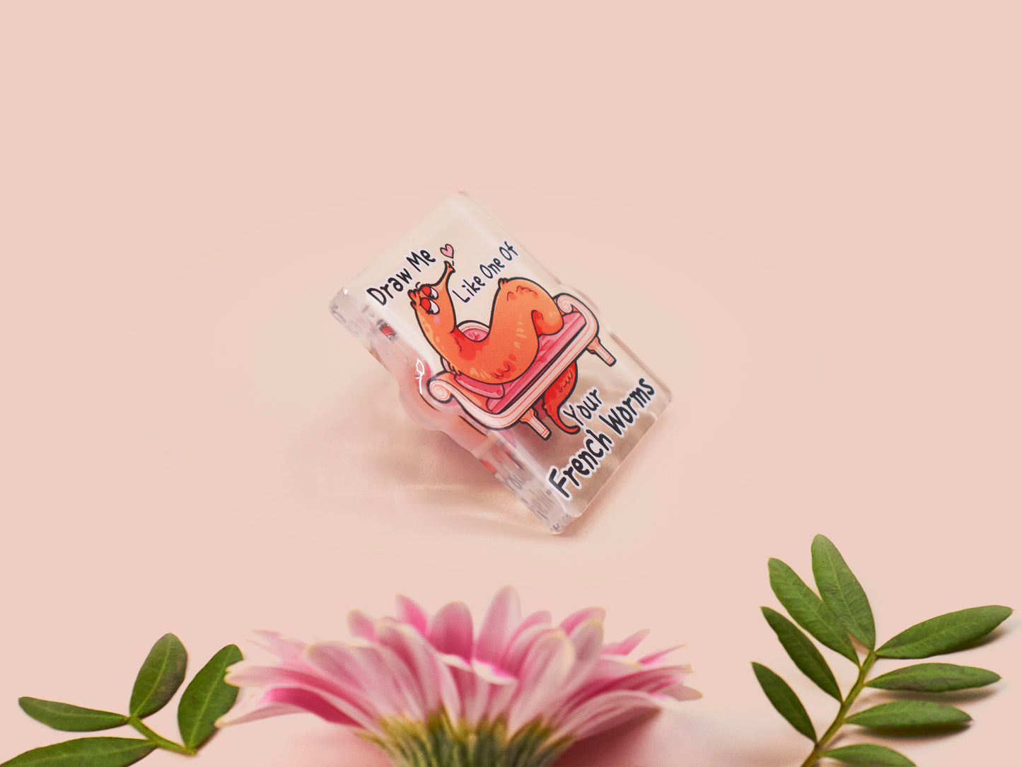 A cute transparent acrylic pin badge with a pink heart-shaped rubber clasp pin back. The design of the pin badge is a cartoon orange worm on a sting sitting on a sofa chaise lounge with the titanic quote Draw Me Like One of Your French Worms