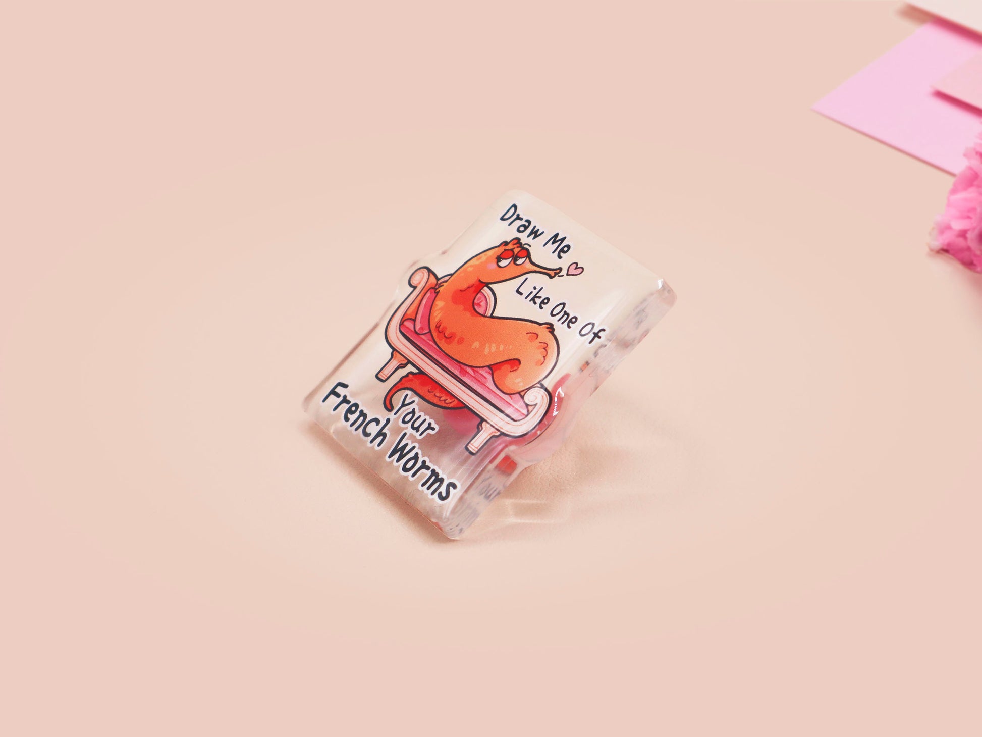 A cute transparent acrylic pin badge with a pink heart-shaped rubber clasp pin back. The design of the pin badge is a cartoon orange worm on a sting sitting on a sofa chaise lounge with the titanic quote Draw Me Like One of Your French Worms