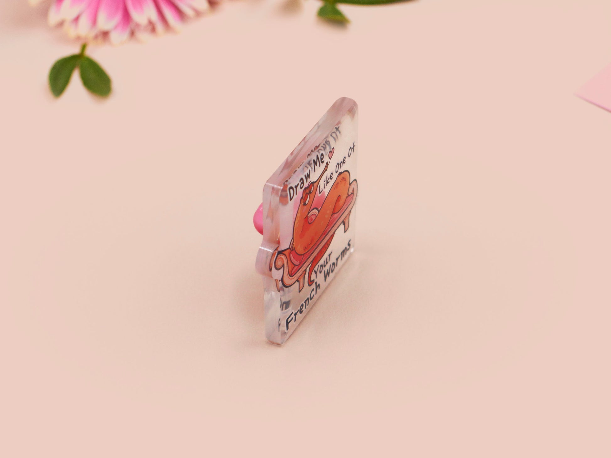 A cute transparent acrylic pin badge with a pink heart-shaped rubber clasp pin back. The design of the pin badge is a cartoon orange worm on a sting sitting on a sofa chaise lounge with the titanic quote Draw Me Like One of Your French Worms