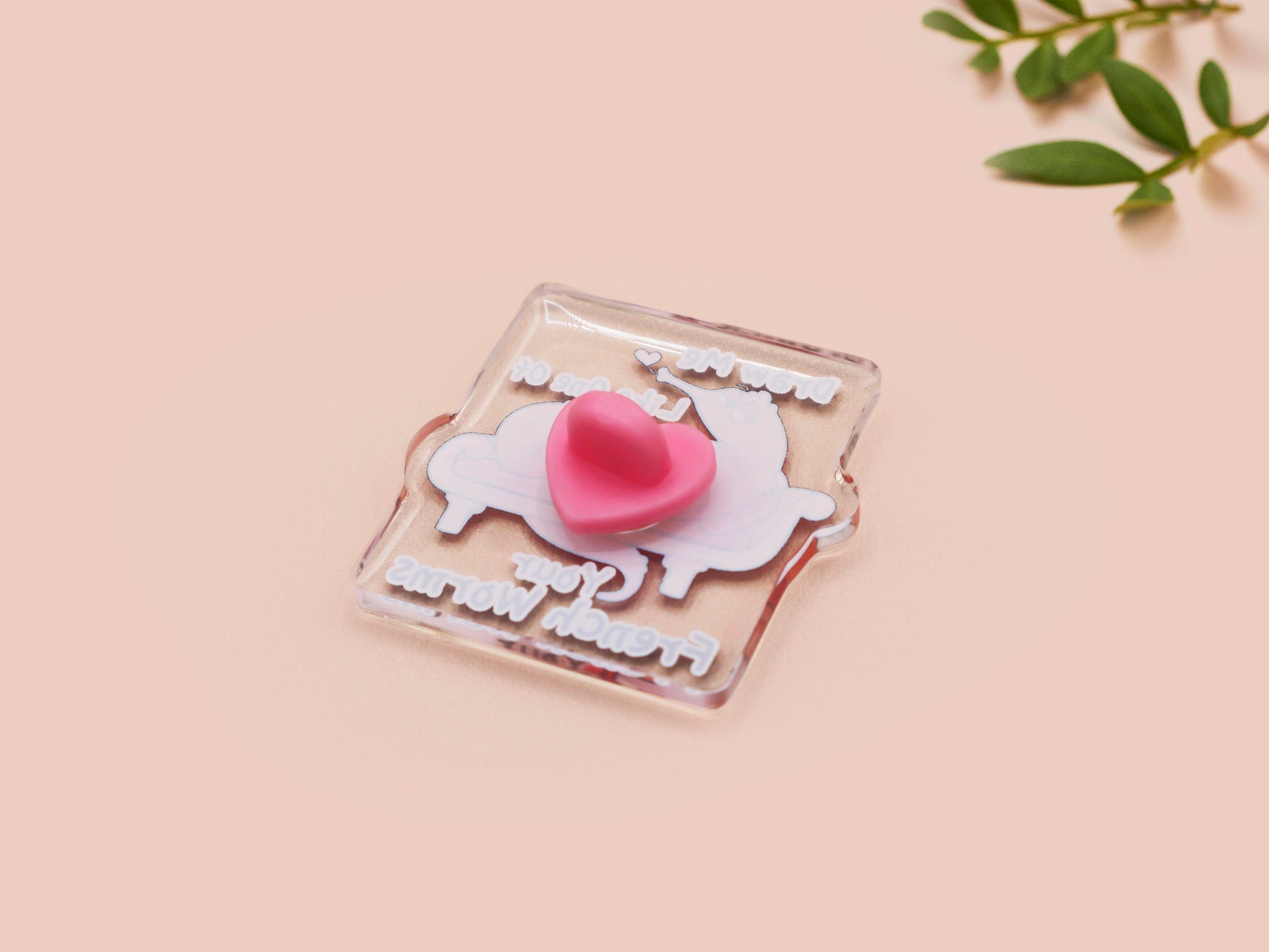 A cute transparent acrylic pin badge with a pink heart-shaped rubber clasp pin back. The design of the pin badge is a cartoon orange worm on a sting sitting on a sofa chaise lounge with the titanic quote Draw Me Like One of Your French Worms