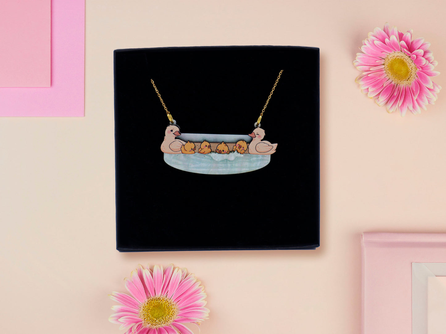 A wooden and glitter acrylic necklace with gold chain in a black box of a cute family of ducks two adult white ducks and four yellow ducklings playing and splashing in the water