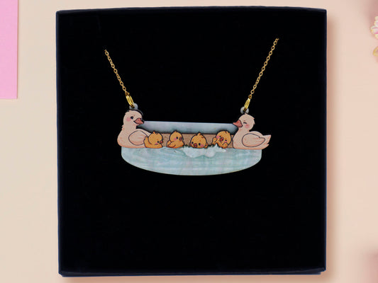 A wooden and glitter acrylic necklace with gold chain in a black box of a cute family of ducks two adult white ducks and four yellow ducklings playing and splashing in the water
