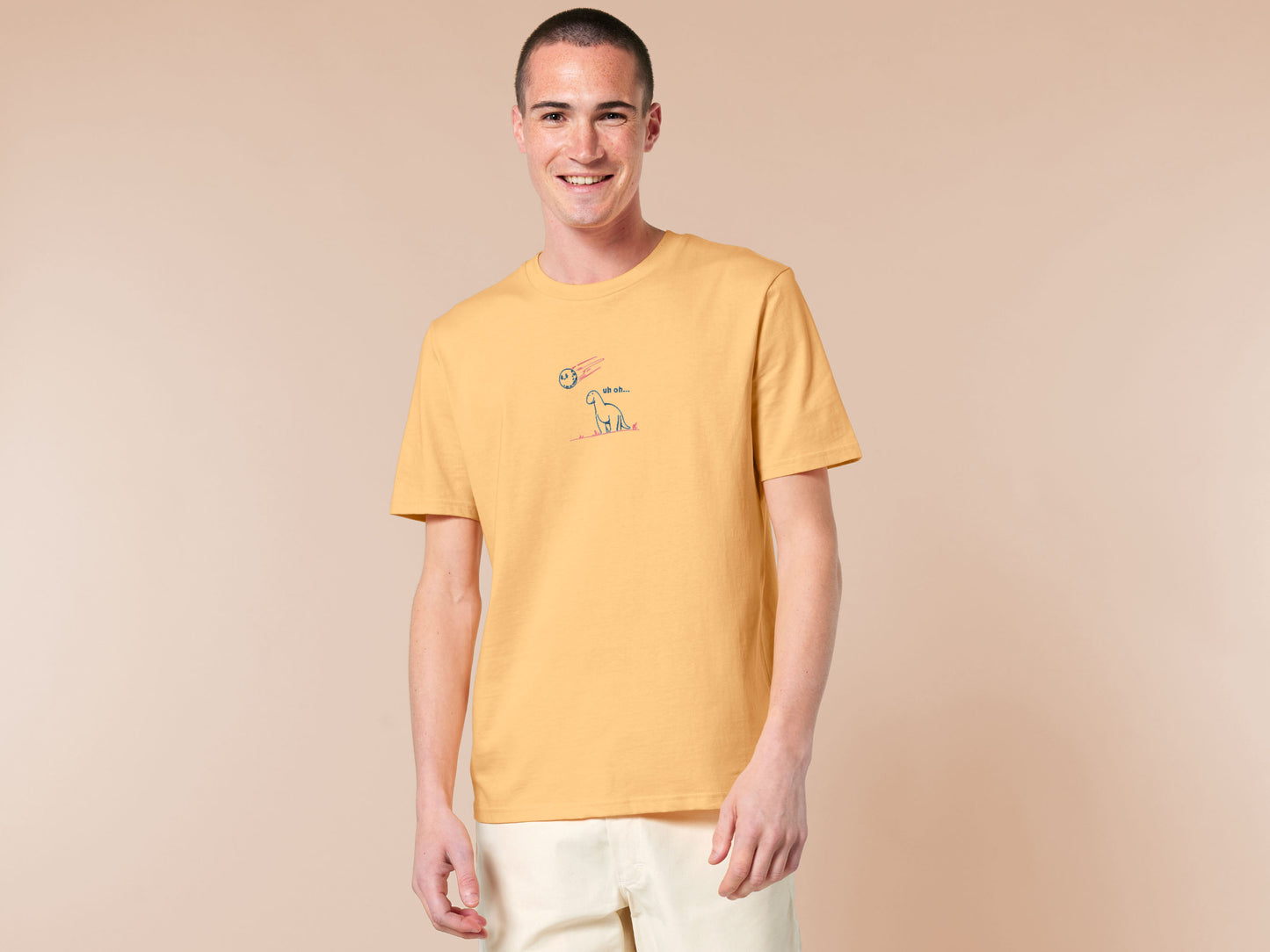 A yellow t-shirt with a blue and pink embroidered design of a cute smiling dinosaur looking at a meteor plummeting to earth with the text uh oh...